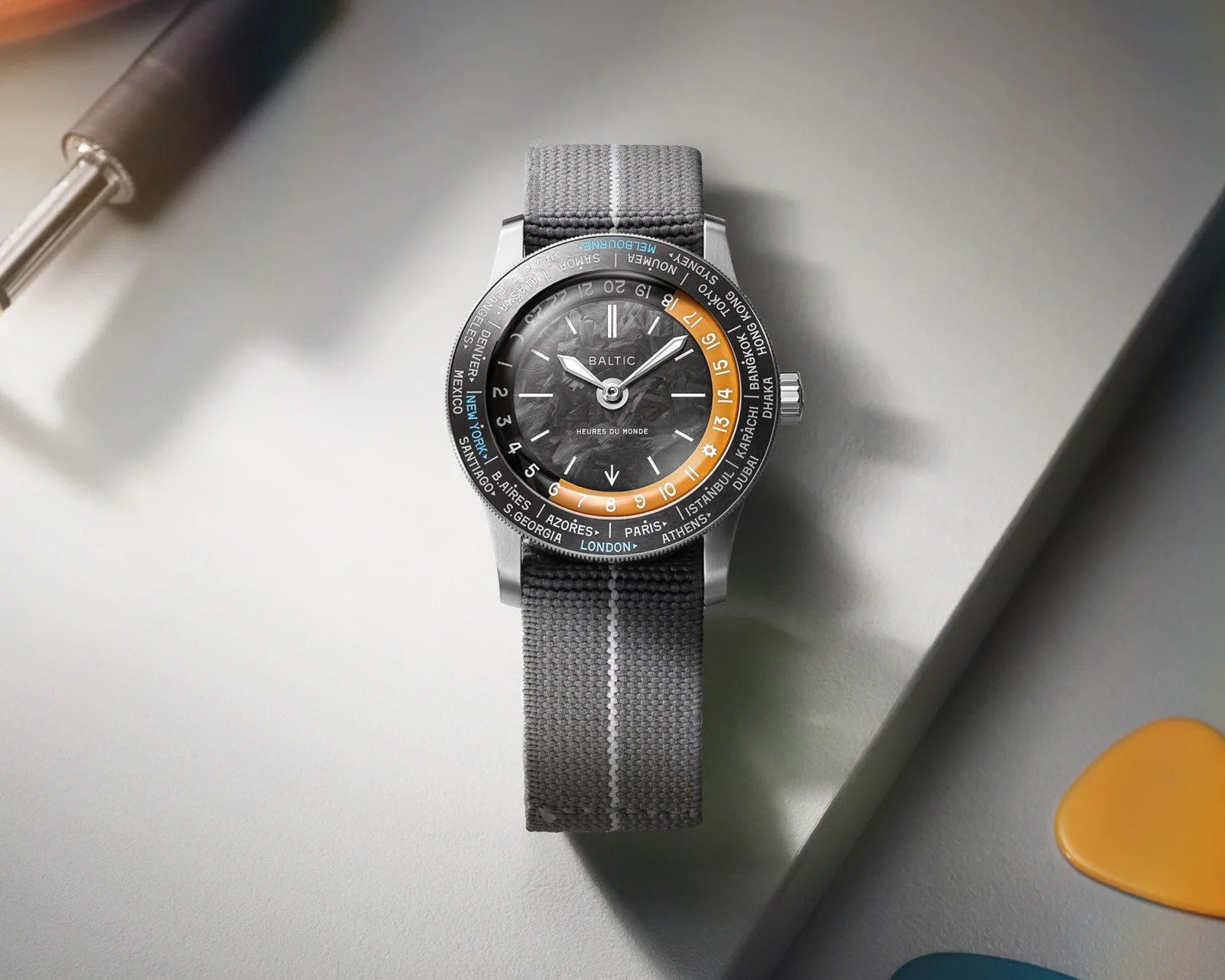Gray fabric strap wristwatch with a black textured dial and orange 24-hour ring, showing world cities on the bezel.