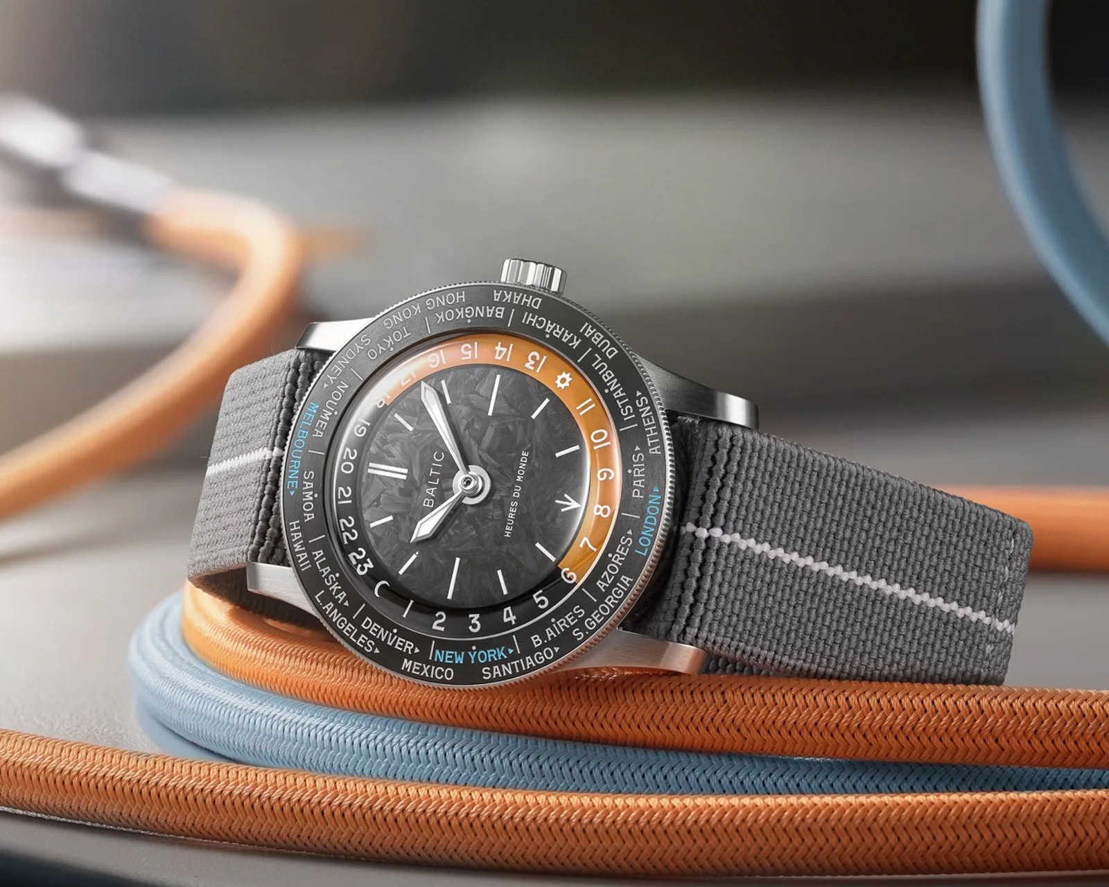 Gray fabric strap wristwatch with black textured dial, orange 24-hour ring, and city names on black bezel.