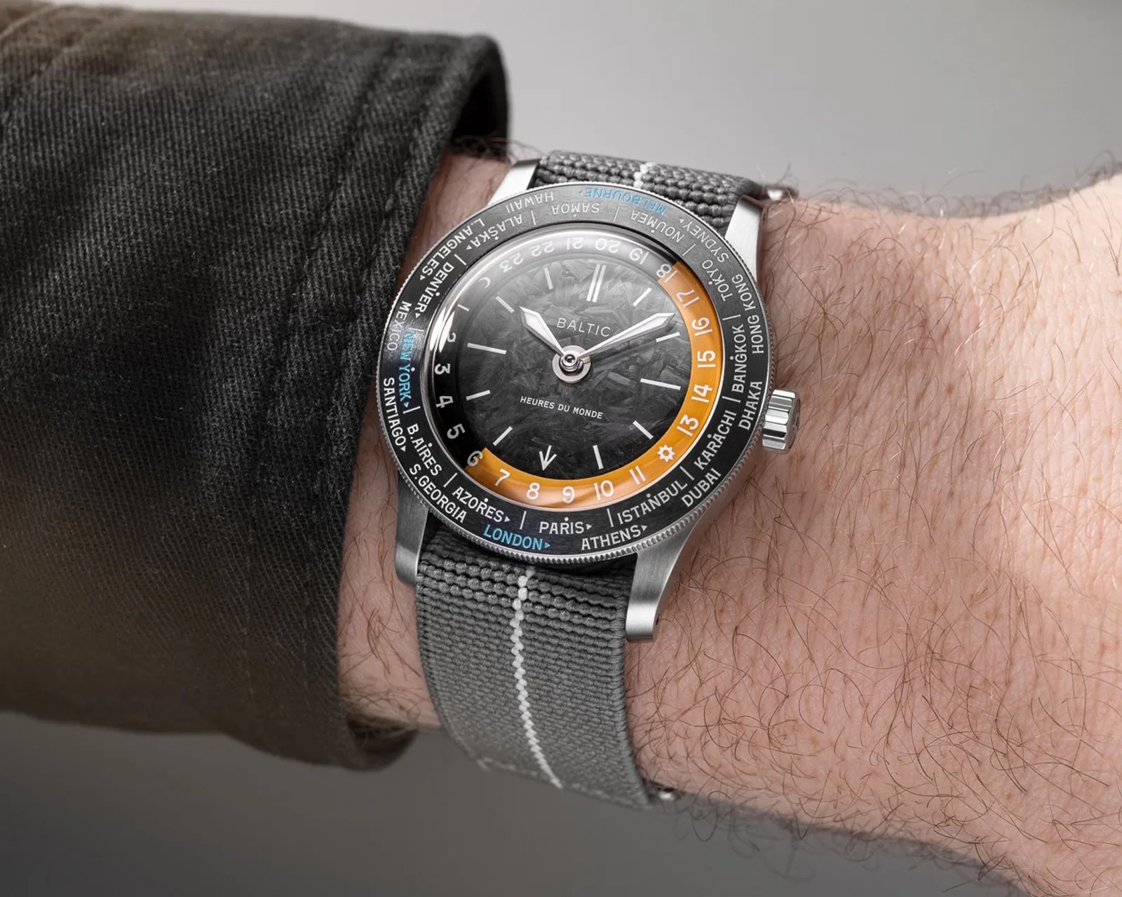 Wristwatch with a textured black dial, orange and black 24-hour ring, and gray fabric strap on a wrist.