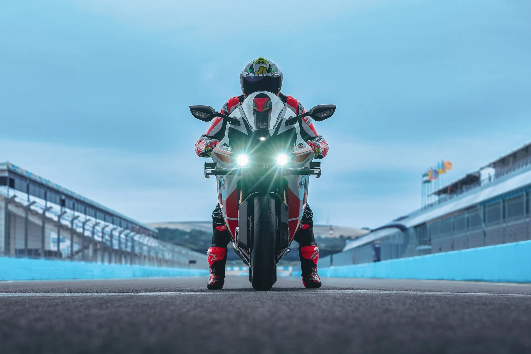 Front view of a white and red sport motorcycle with headlights on, ridden by a person in matching gear on a race track.