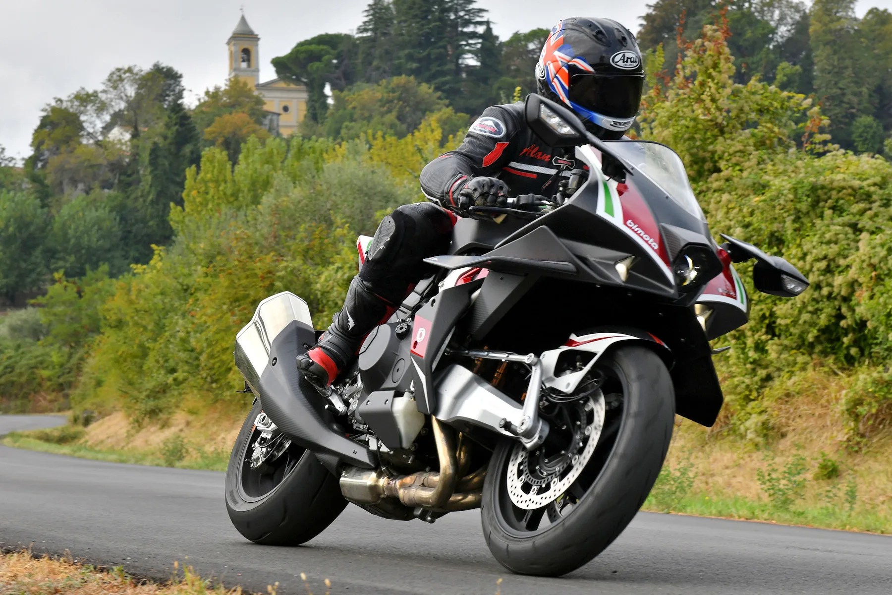 Black and red Bimota sport motorcycle leaning into a turn on a road with green trees and a church tower in the background.