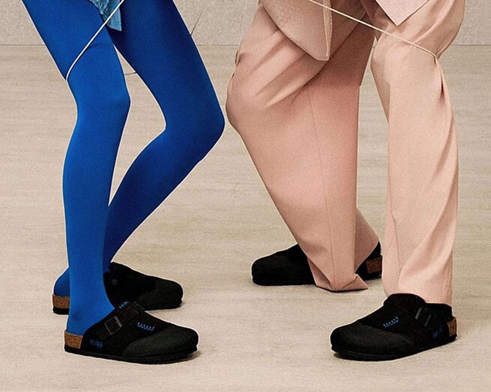 Two pairs of legs wearing black shoes, one in blue leggings and the other in beige pants, standing on a light floor.