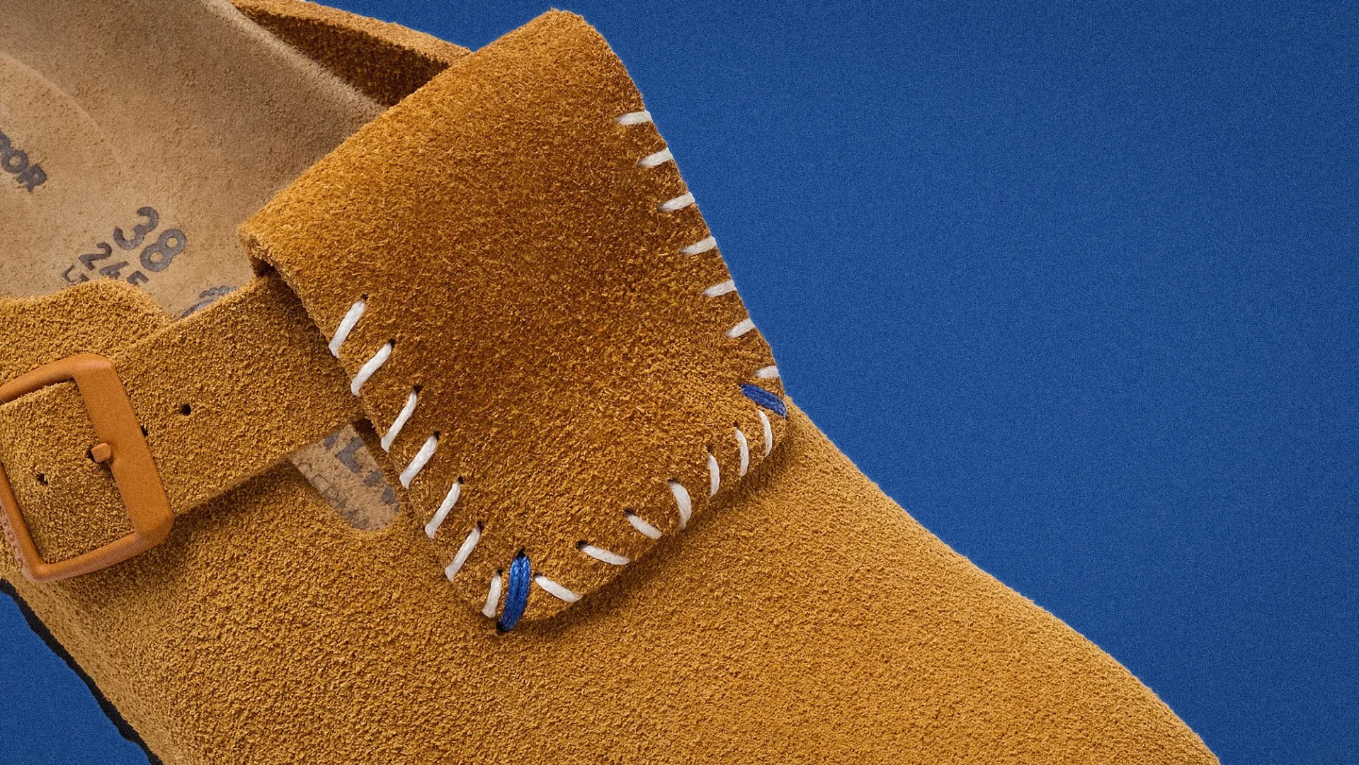 Close-up of a tan suede shoe with white and blue stitching and a tan buckle on a blue background.