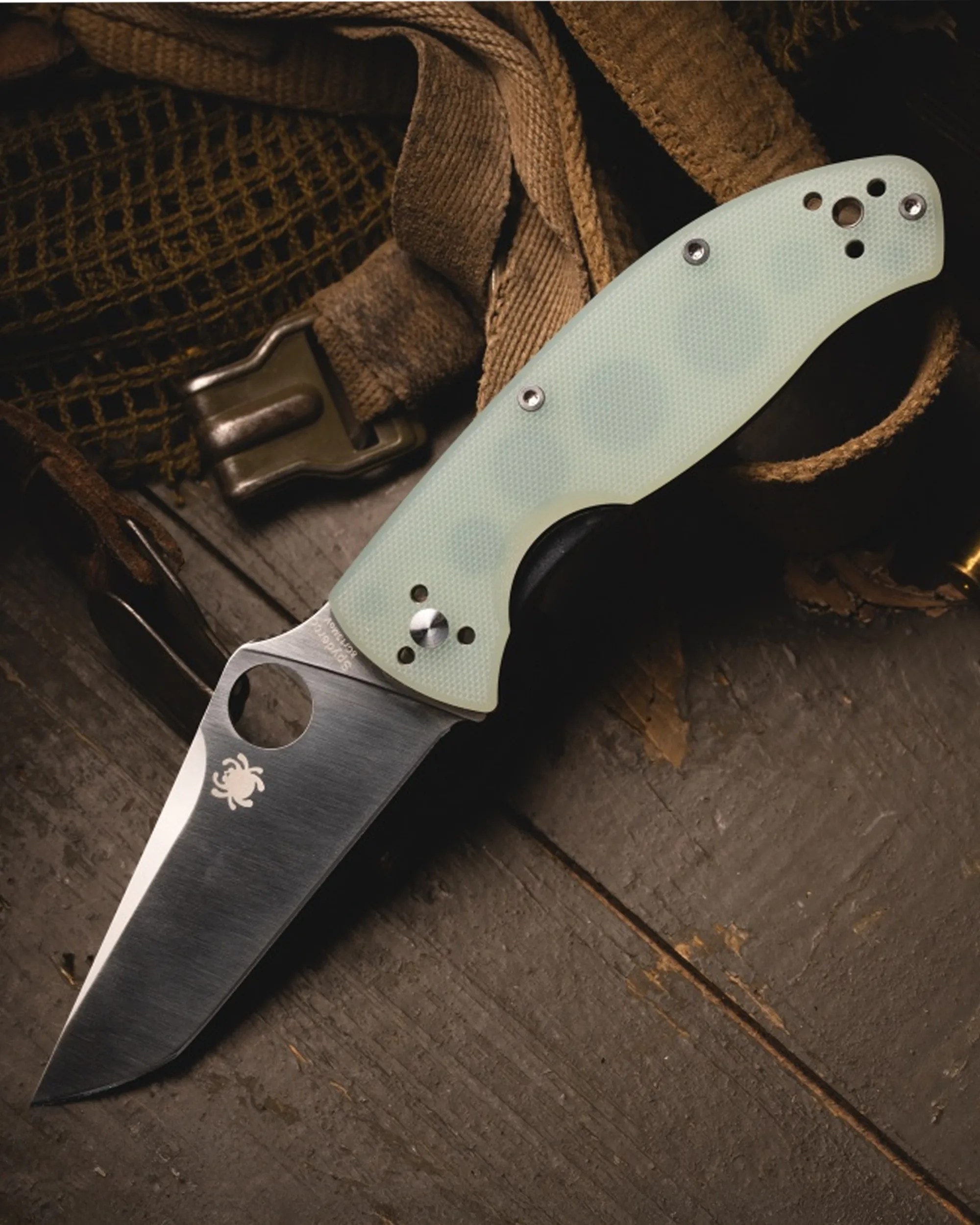 Folding knife with a light green textured handle and a stainless steel blade resting on a wooden surface with brown straps.