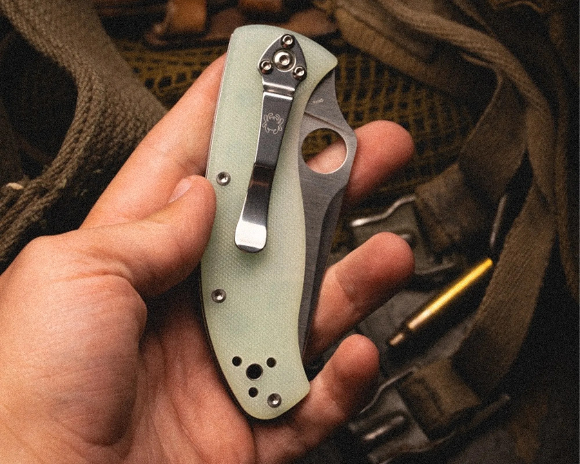 Hand holding a closed folding knife with a light green textured handle and a metal clip with a spider logo.