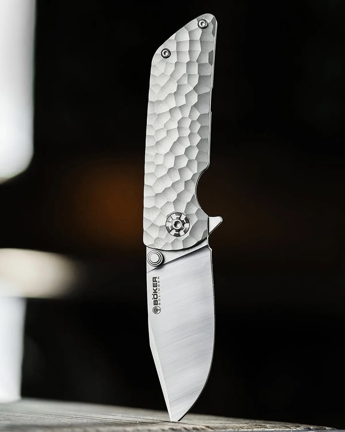 Silver folding knife with a textured handle and Böker logo on the blade standing on a wooden surface.