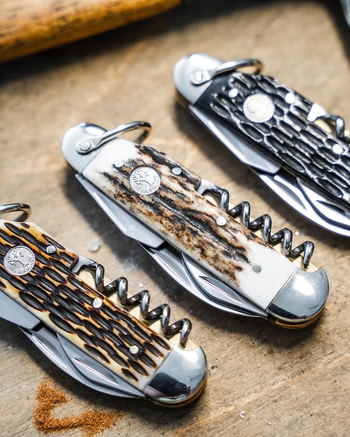 Three closed pocket knives with textured handles in black, brown, and white on a wooden surface.