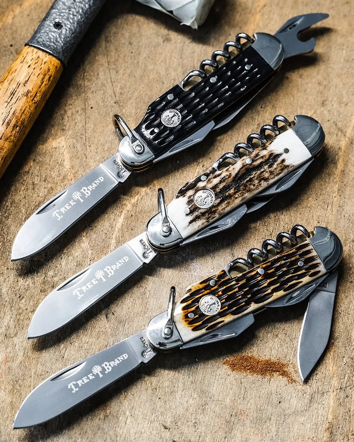 Three Tree Brand folding pocket knives with corkscrews and bottle openers, featuring black, white, and brown textured handles on a wooden surface.