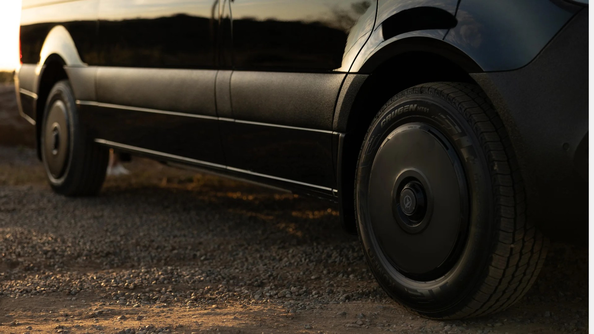 Close-up of black vehicle's side panel and rear wheel with Crugen HT51 tire on gravel ground.