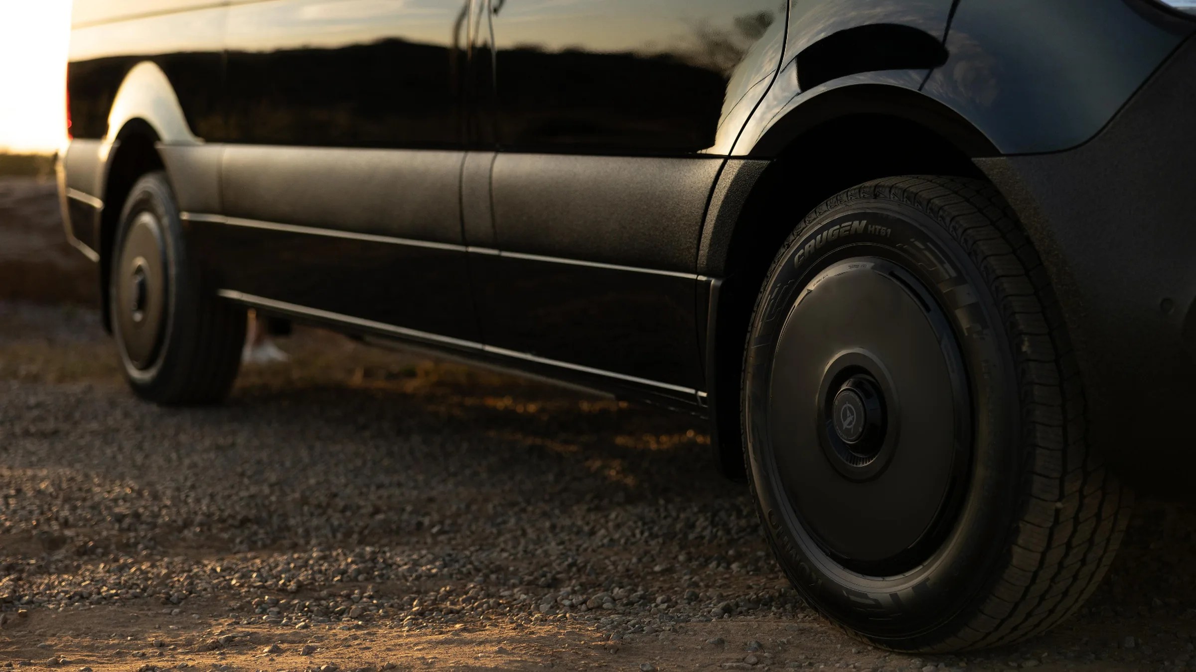 Close-up of a black vehicle's side panel and wheel with a matte black hubcap on a gravel surface at sunset.