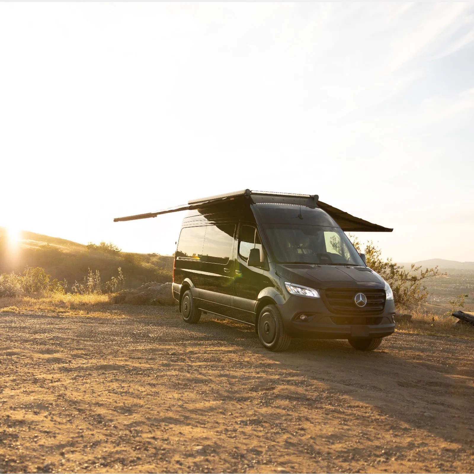 Black Mercedes-Benz camper van with extended side awnings parked on a dirt road at sunset.