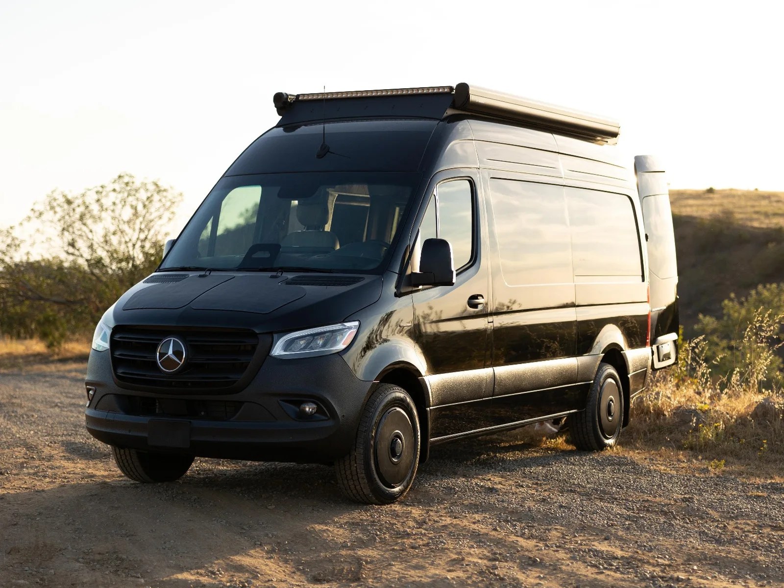 Black Mercedes-Benz camper van with roof light bar parked on a dirt road at sunset.