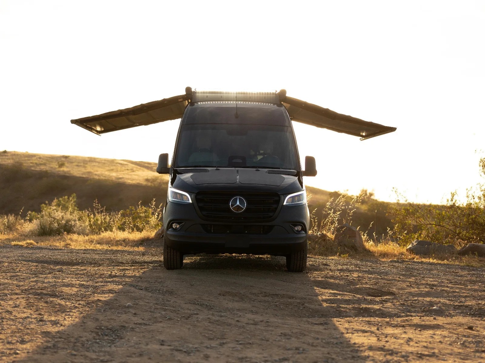Black Mercedes-Benz van with extended side awnings parked on a dirt road at sunset.