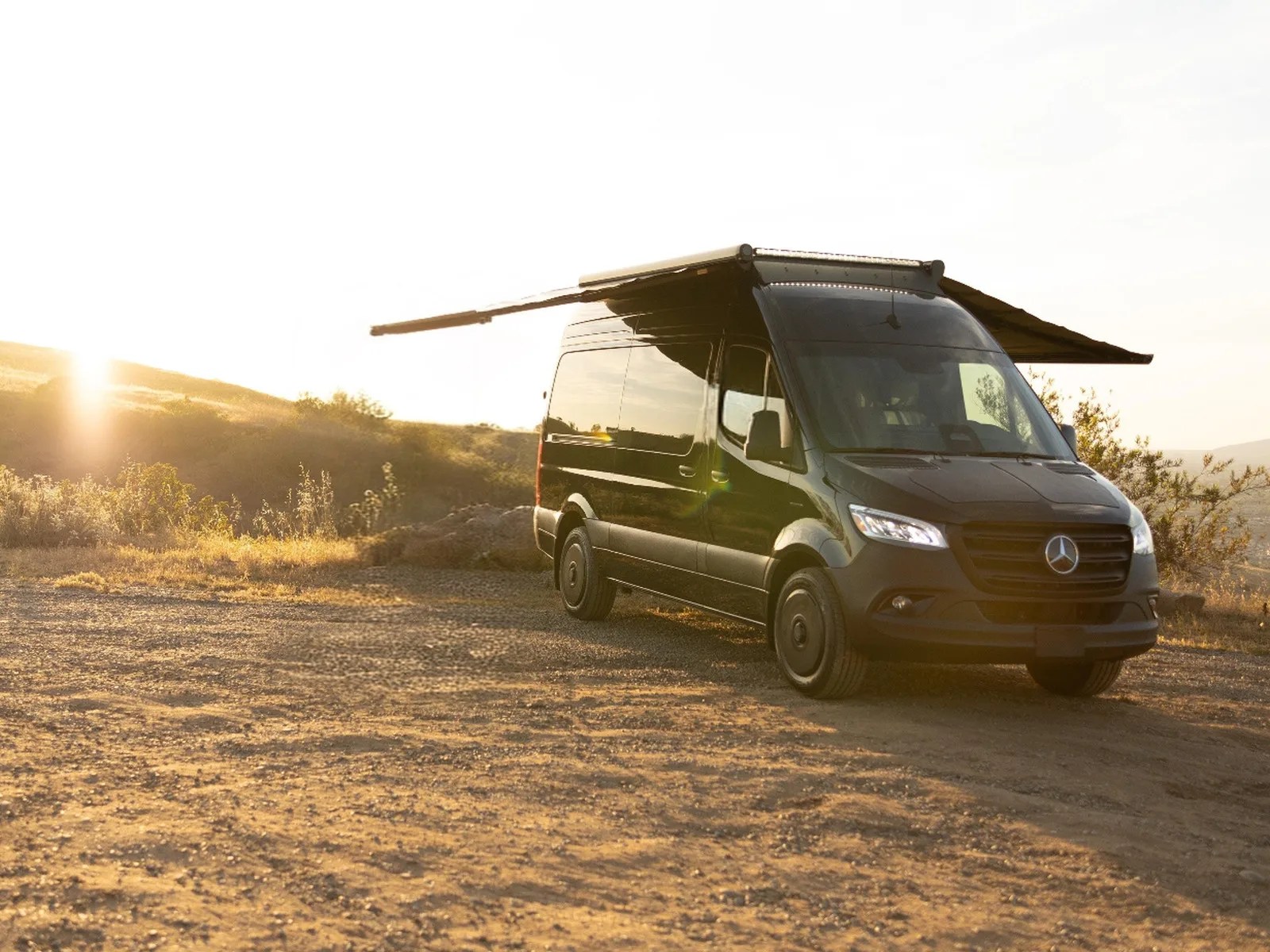 Black Mercedes-Benz camper van with extended side awnings parked on a dirt road at sunset.