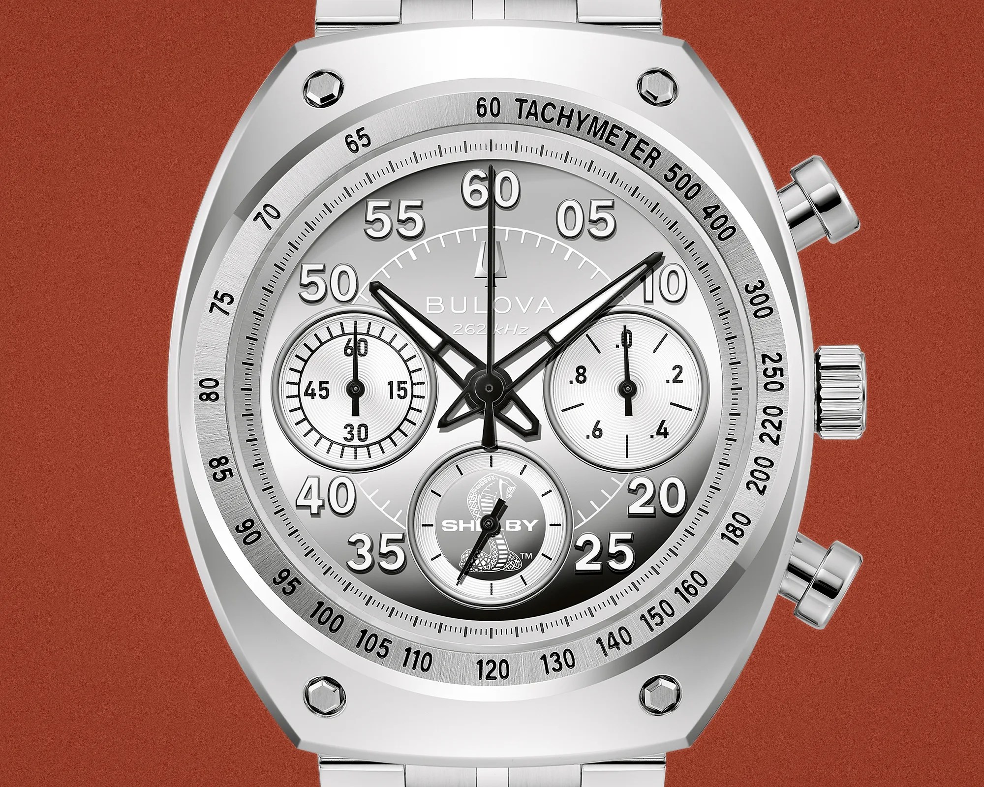 Close-up of a silver Bulova chronograph watch face with tachymeter scale and Shelby cobra logo on a rust-colored background.