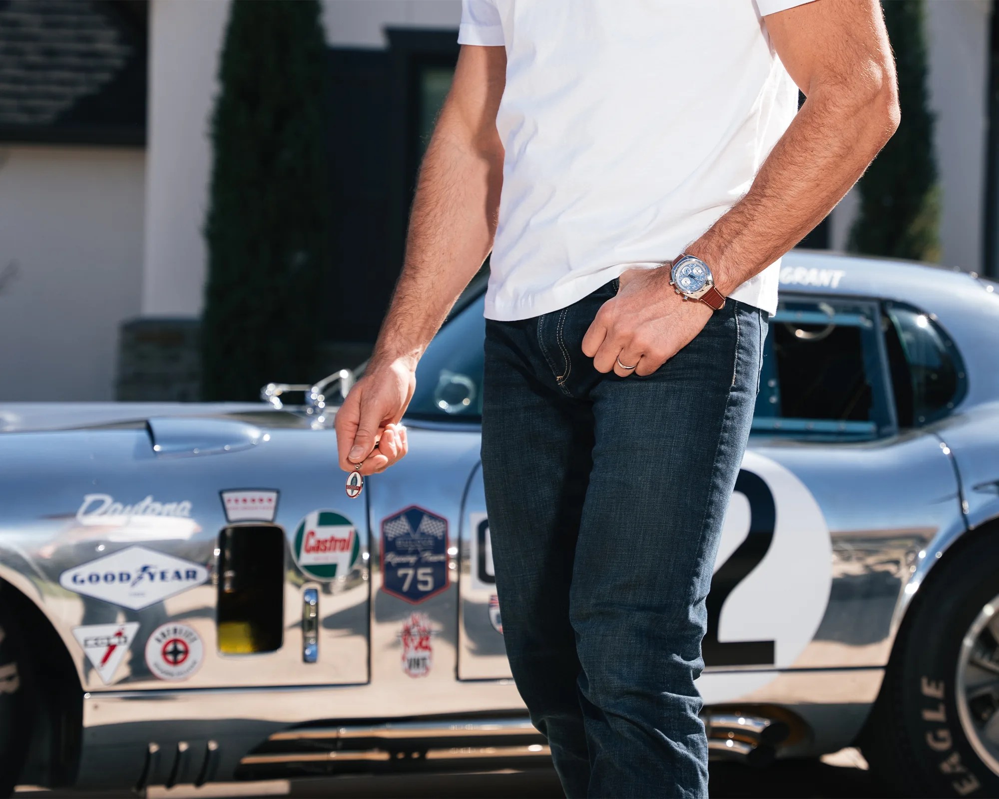 Person wearing a white t-shirt and blue jeans holding a keychain with a silver vintage race car in the background.