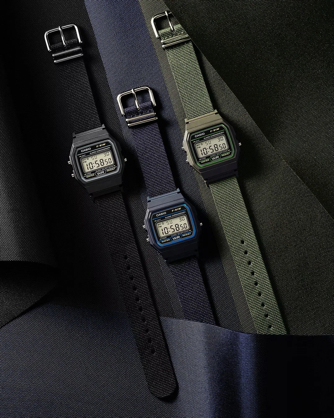Three Casio F-91W digital watches with black, navy blue, and olive green nylon straps on textured fabric.