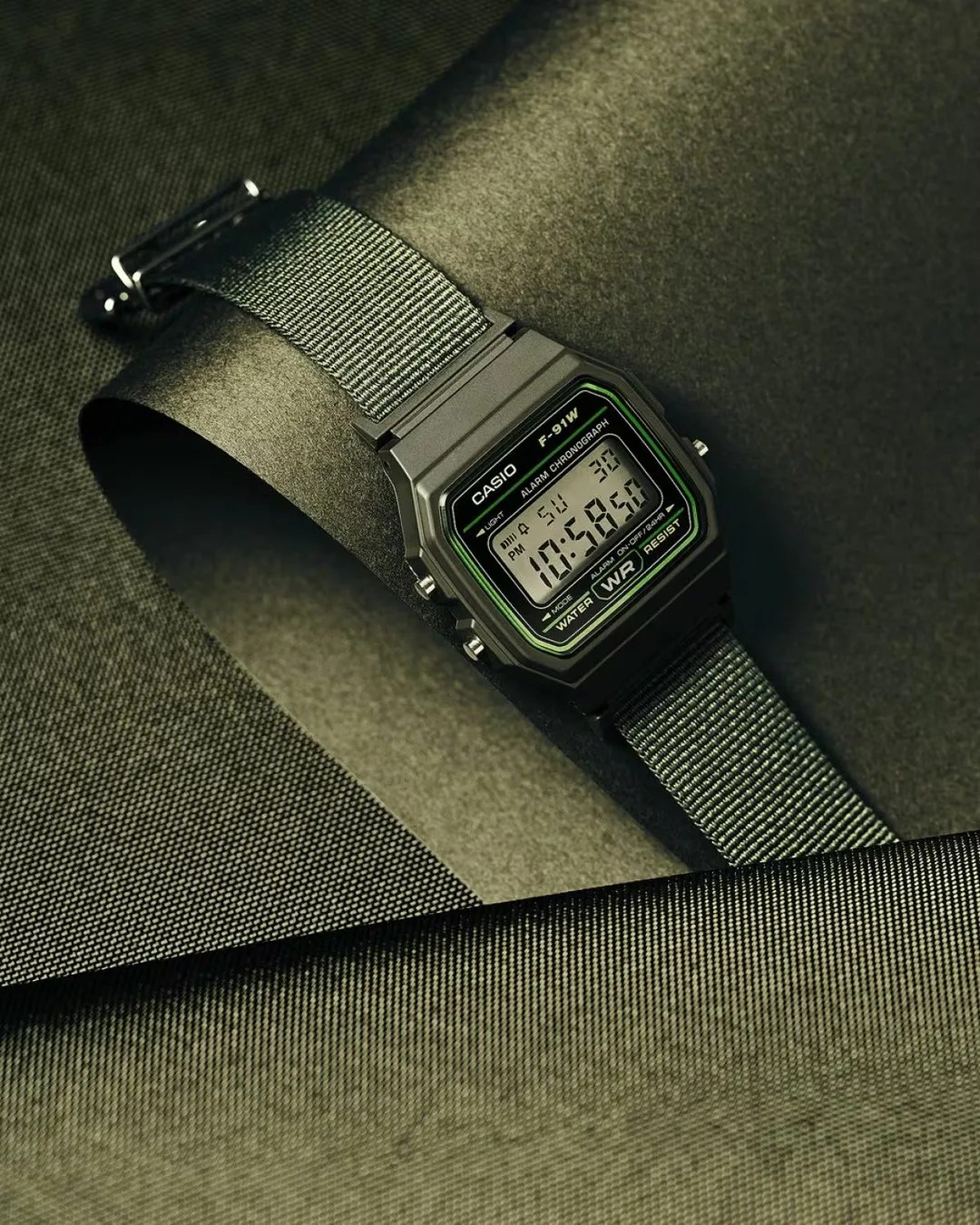 Black Casio F-91W digital watch with green accents and a black nylon strap on textured dark fabric.