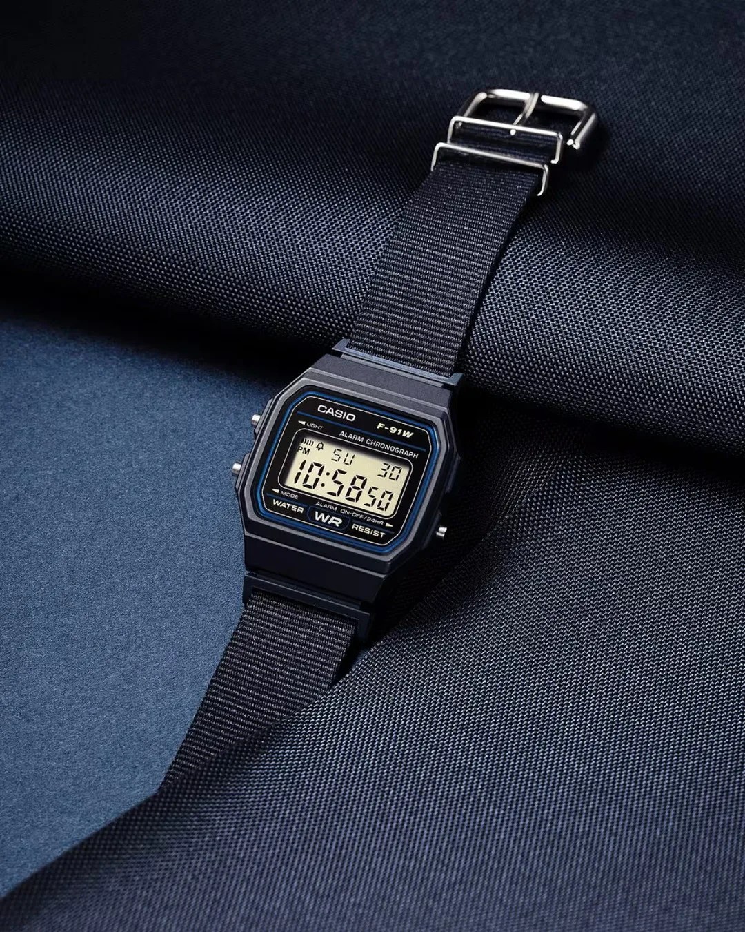 Black Casio F-91W digital watch with black nylon strap on dark textured fabric.