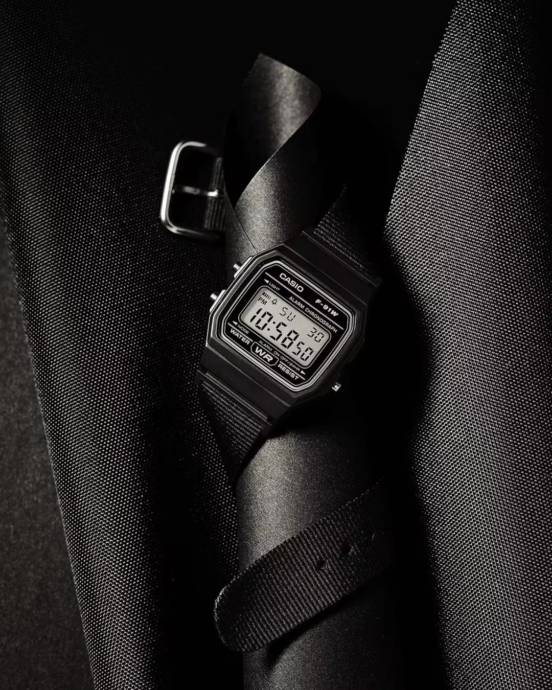 Black Casio F-91W digital watch with a black fabric strap wrapped around a black textured fabric roll.