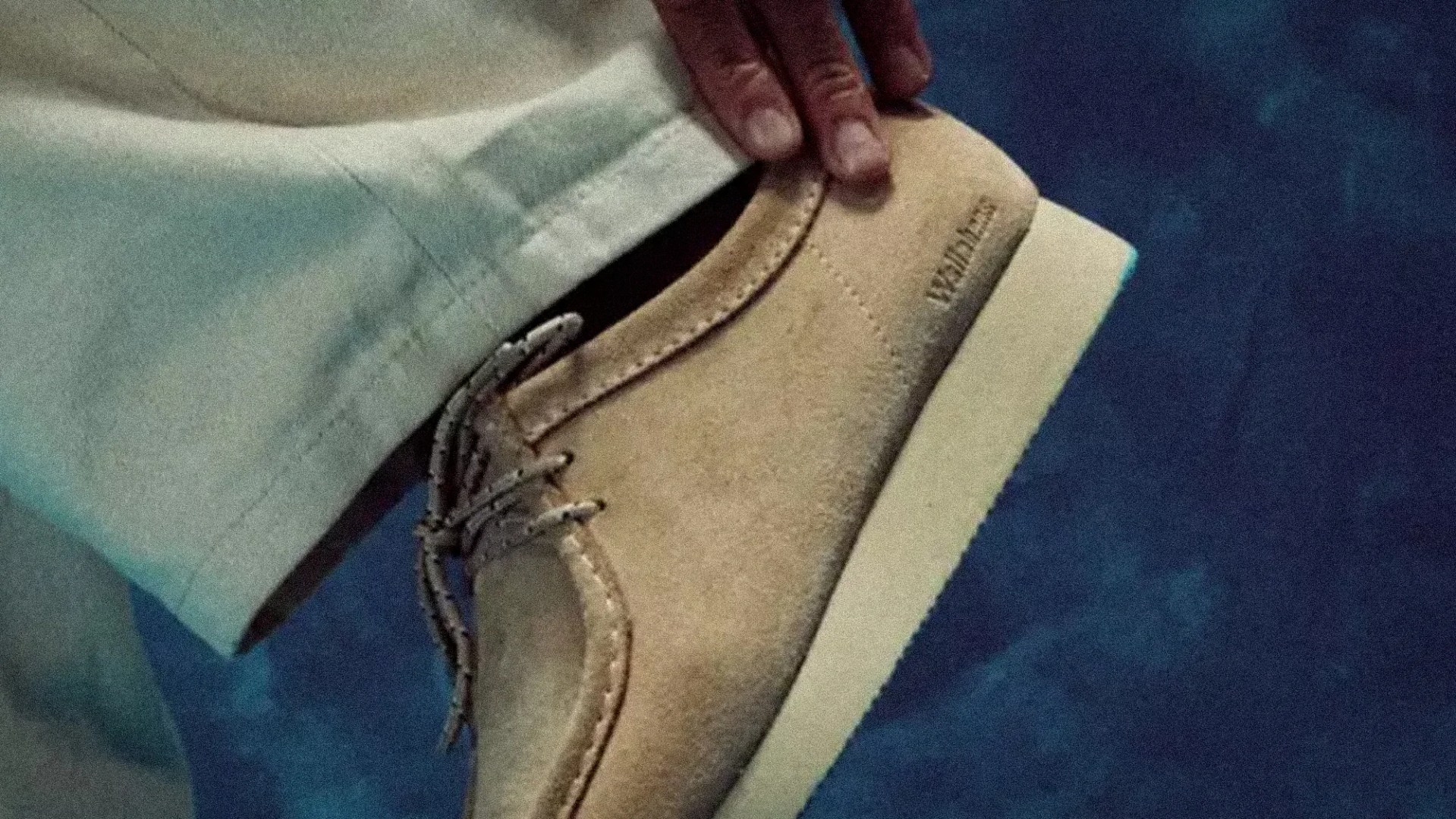 Beige suede Wallabee shoe with white sole and patterned laces worn with light-colored pants.