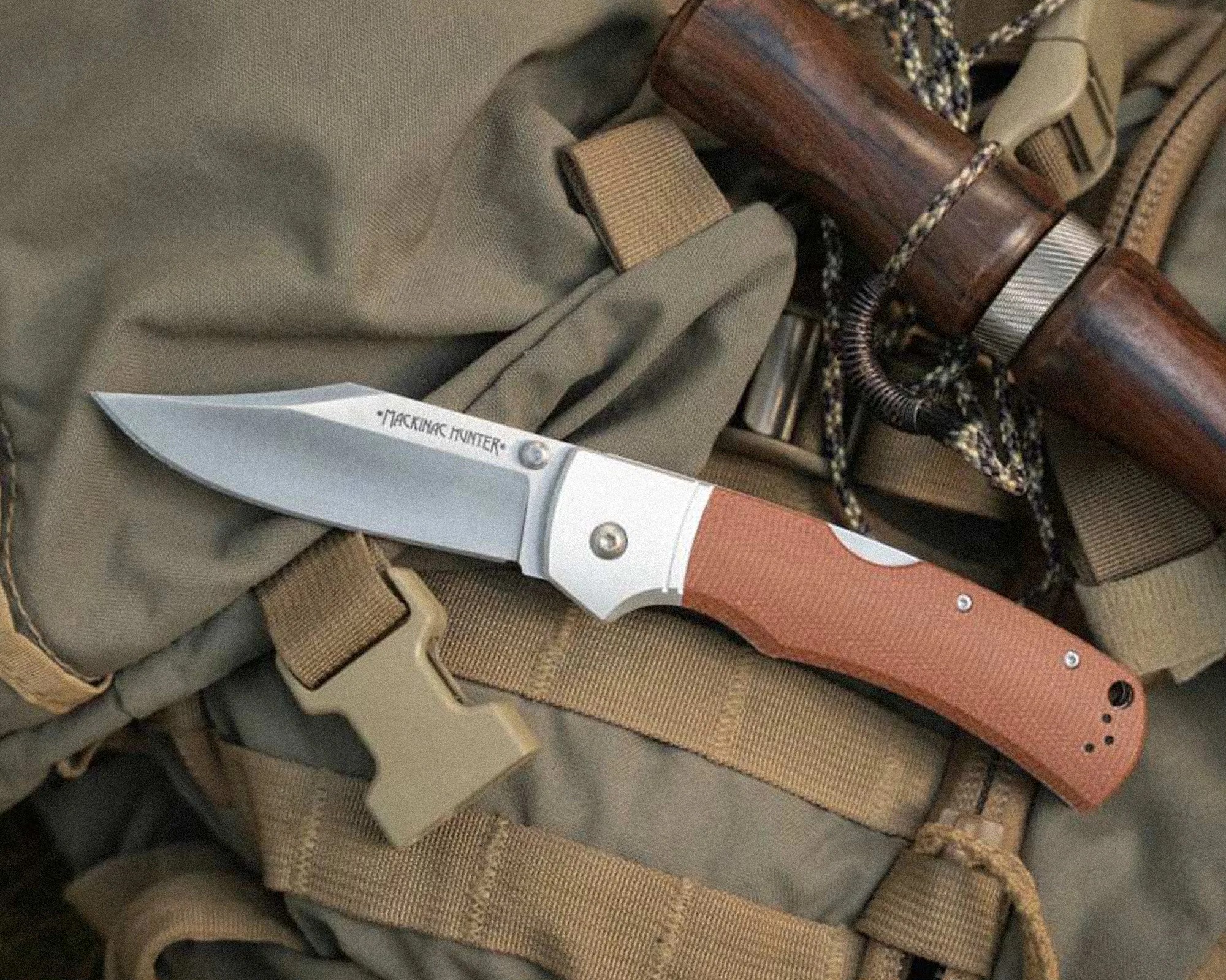 Folding knife with a silver blade and brown textured handle resting on tan tactical gear with straps and buckles.