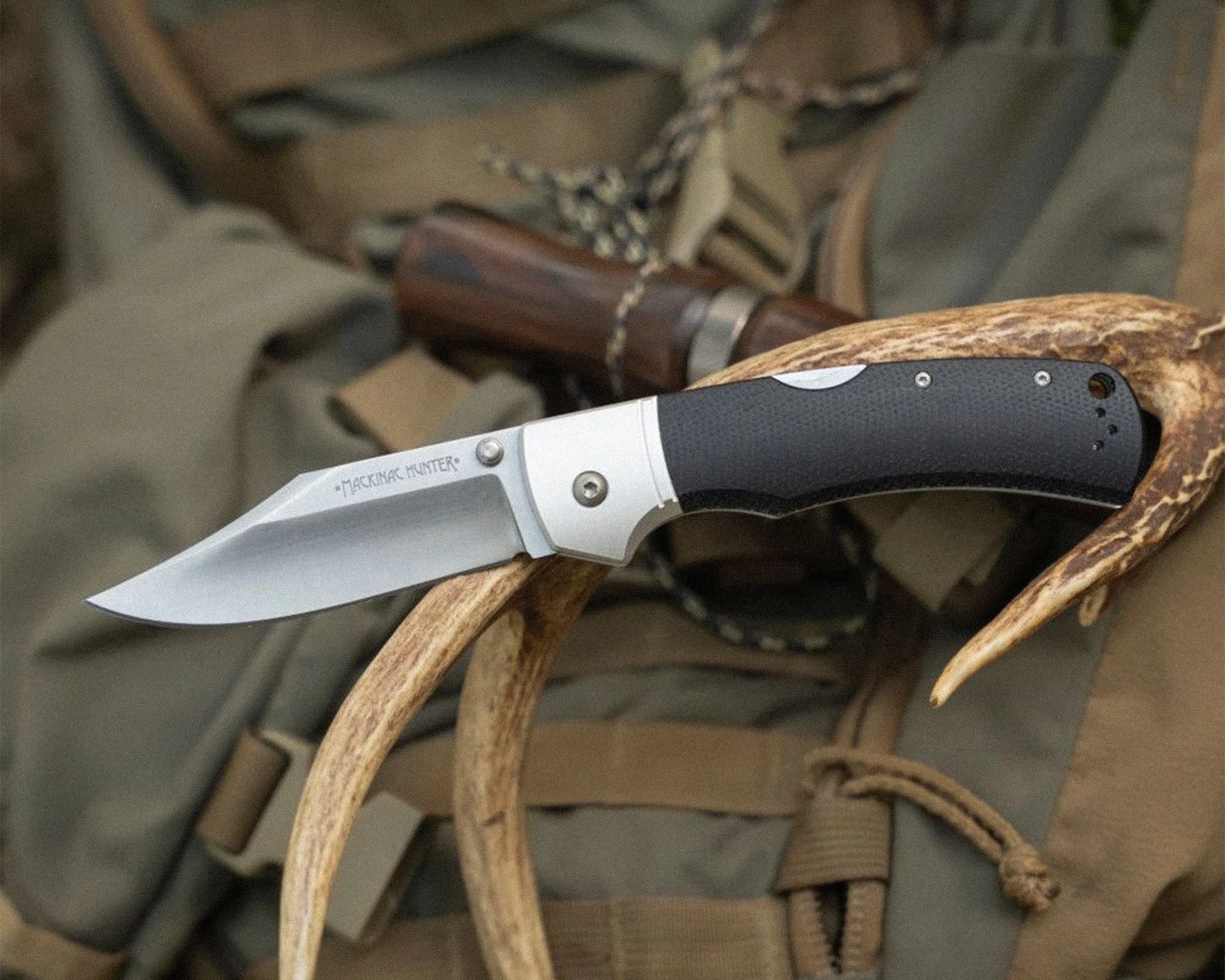 Folding hunting knife with black textured handle and silver blade resting on antlers and outdoor gear.