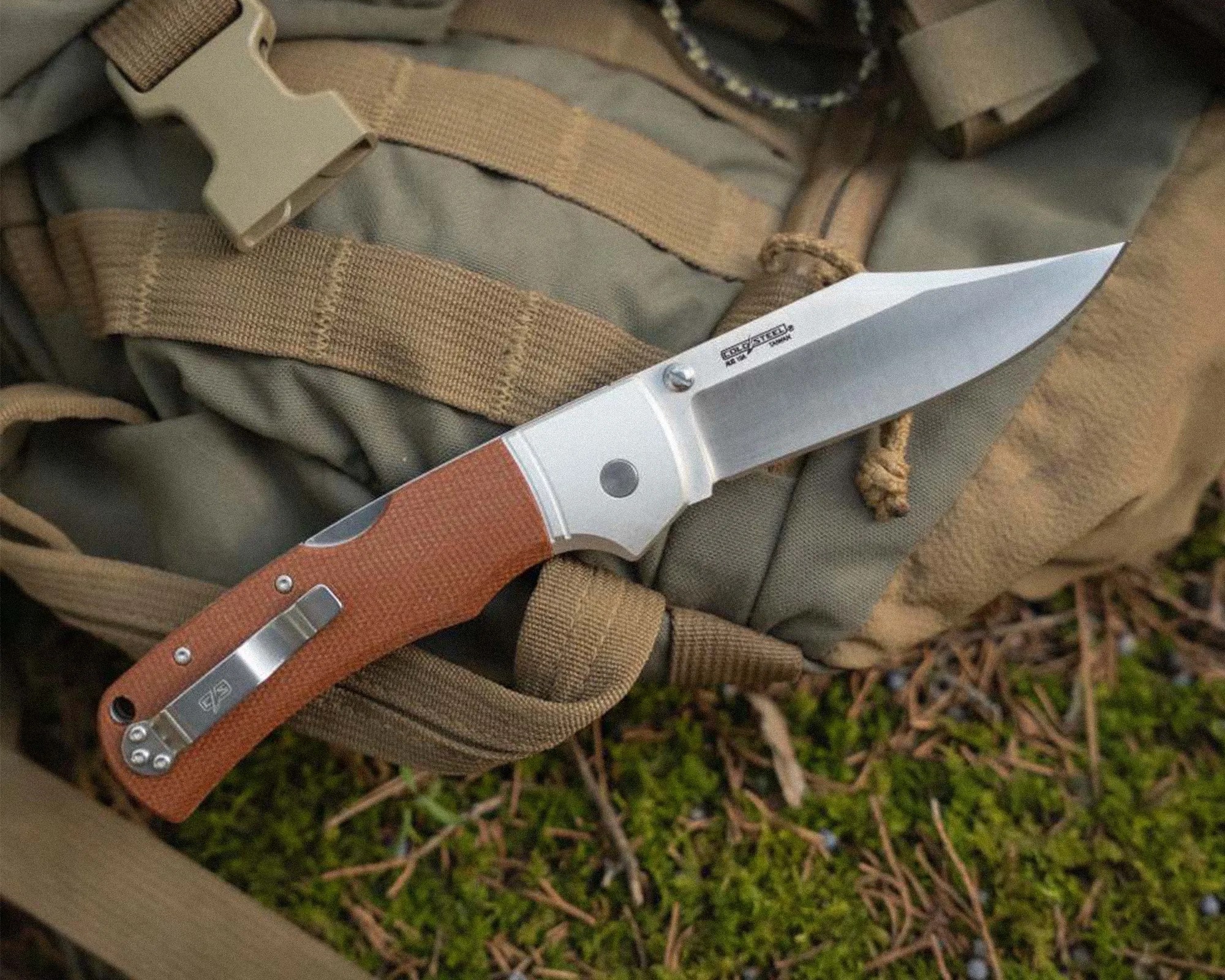 Folding knife with a brown textured handle and stainless steel blade resting on green and tan fabric with straps.