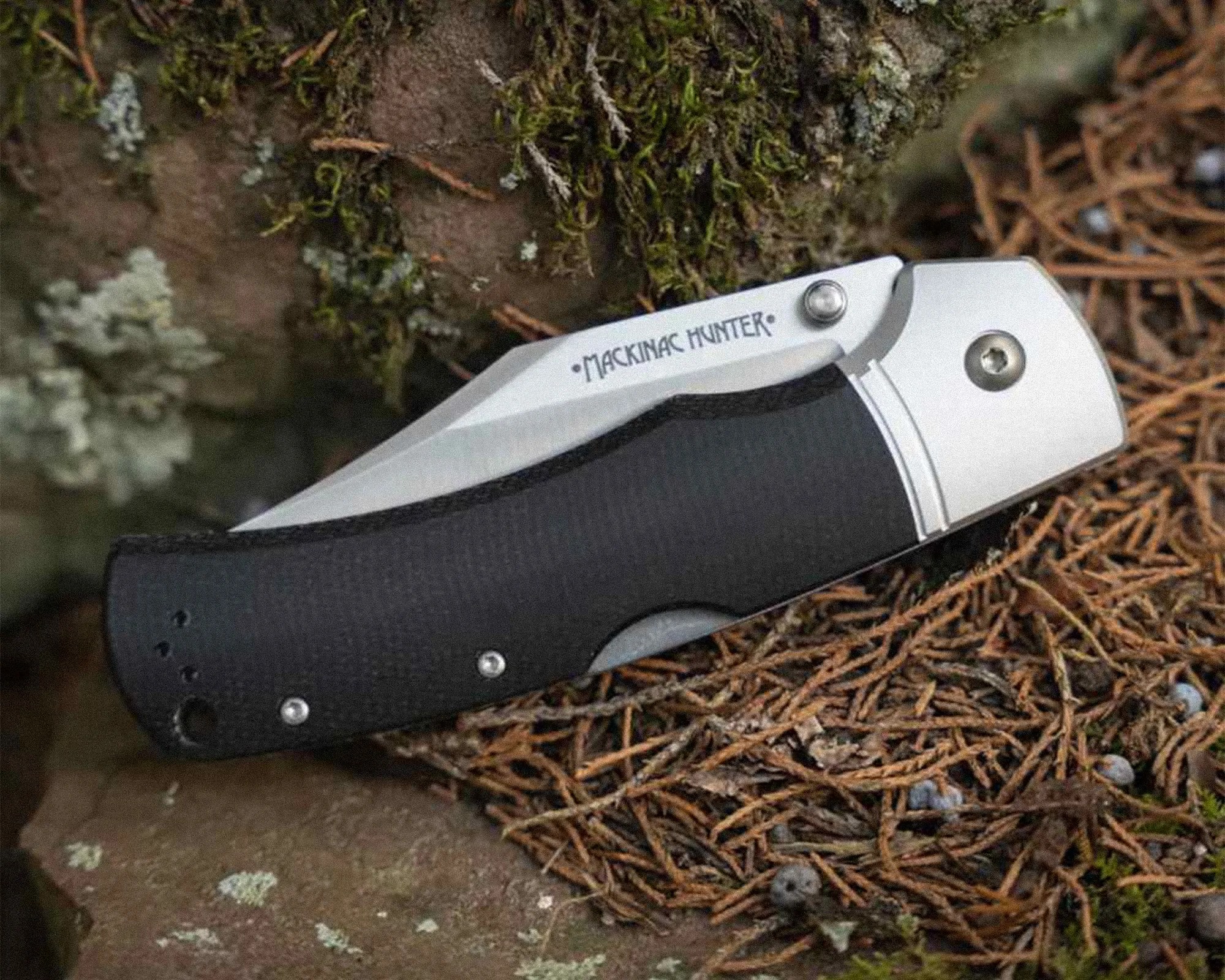 Folding knife with a black textured handle and a partially visible blade engraved with "MACKINAC HUNTER" resting on mossy ground.