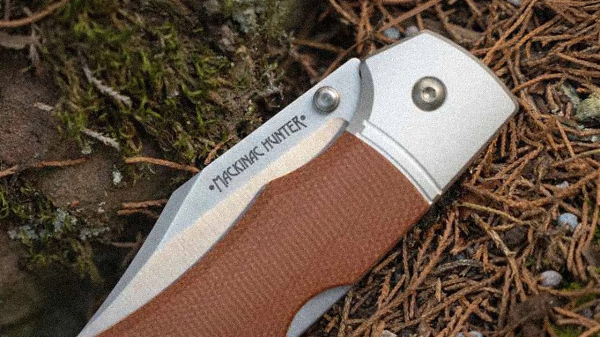 Close-up of a folding knife blade with "MACKINAC HUNTER" engraved, featuring a brown textured handle and metal bolsters.