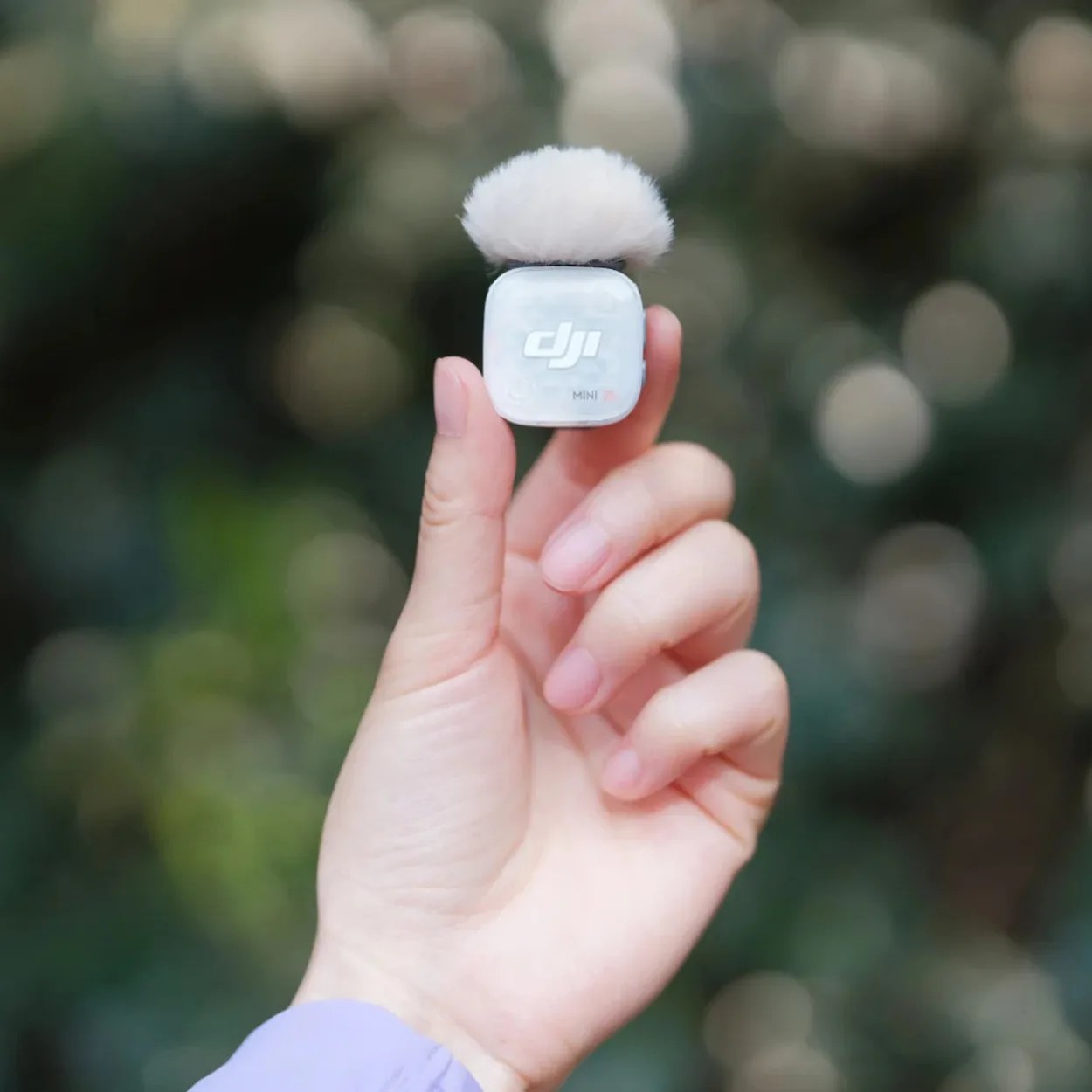 Hand holding a small white DJI Mini 3 microphone with a fluffy windscreen against a blurred outdoor background.