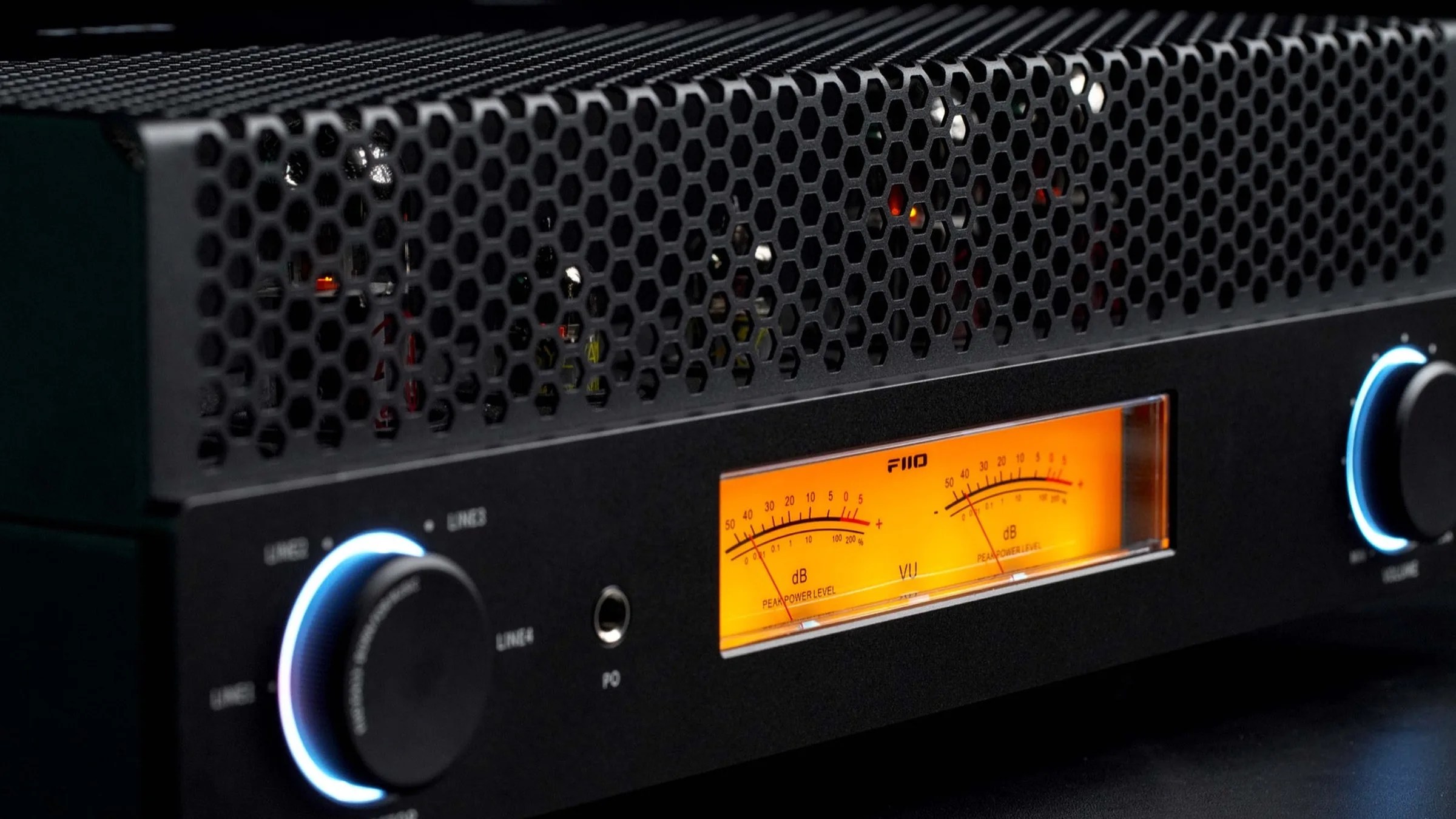 Black audio amplifier with hexagonal ventilation grill, two illuminated control knobs, and orange VU meter display.