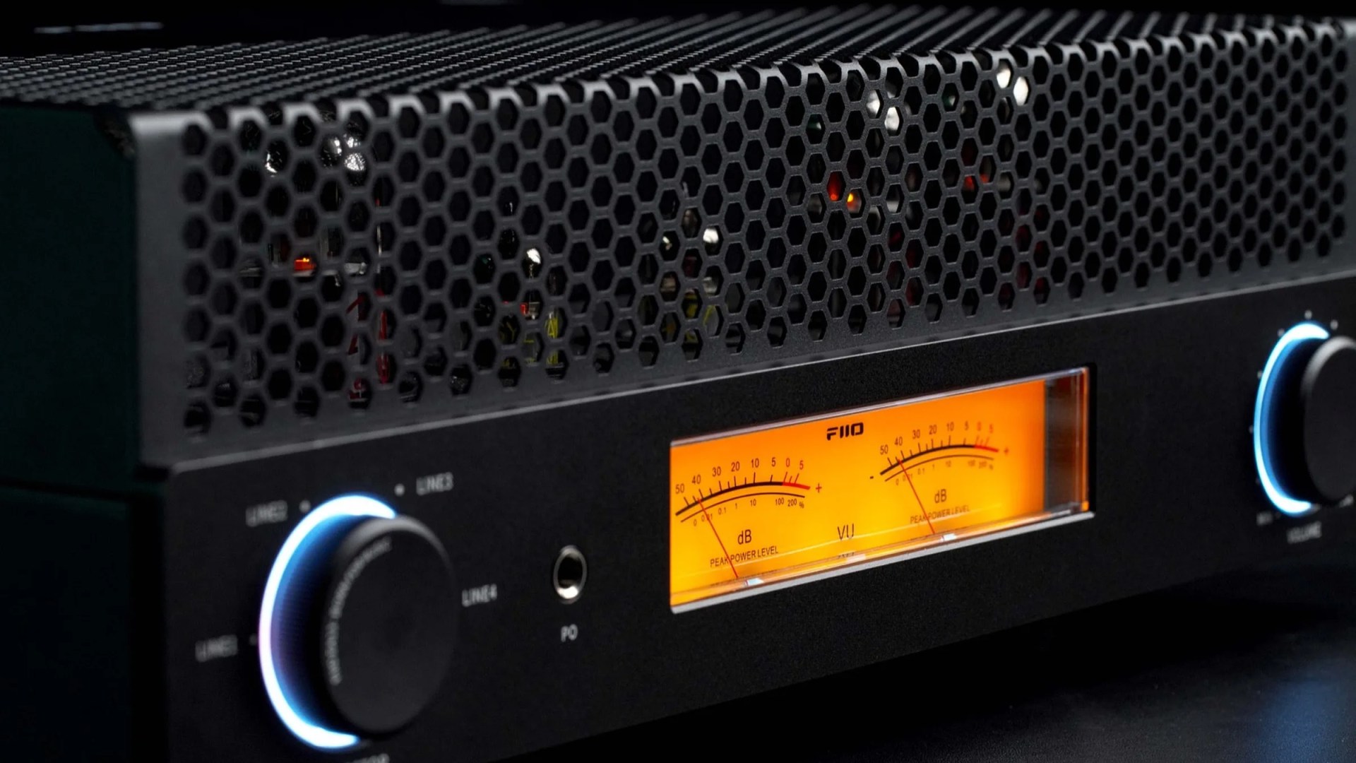 Black audio amplifier with hexagonal ventilation grill, two illuminated control knobs, and orange VU meter display.