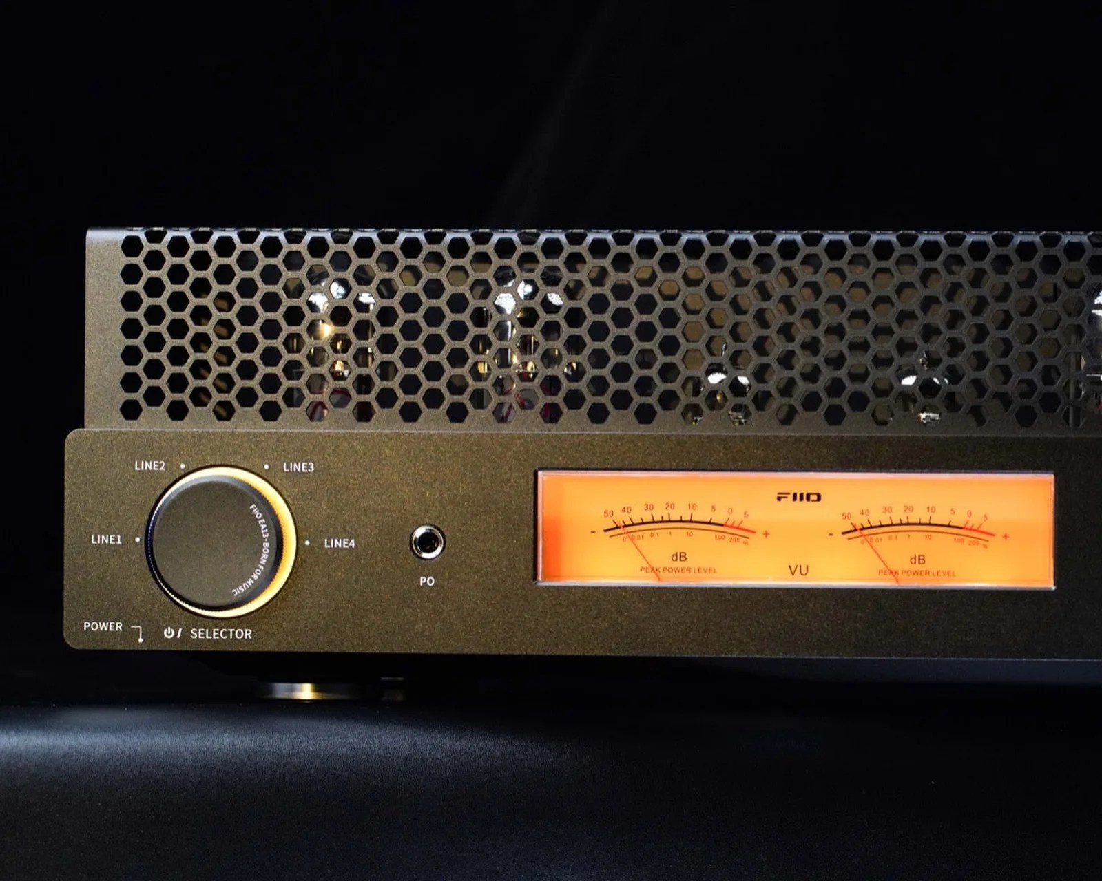 Black audio amplifier with hexagonal ventilation grill, large selector knob, and orange VU meters.