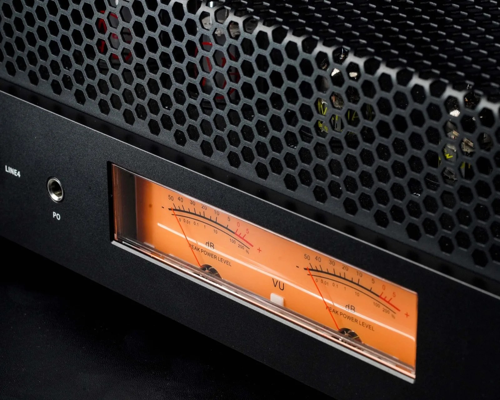 Close-up of a black audio amplifier with a honeycomb metal grille and an orange VU meter display showing peak power levels.