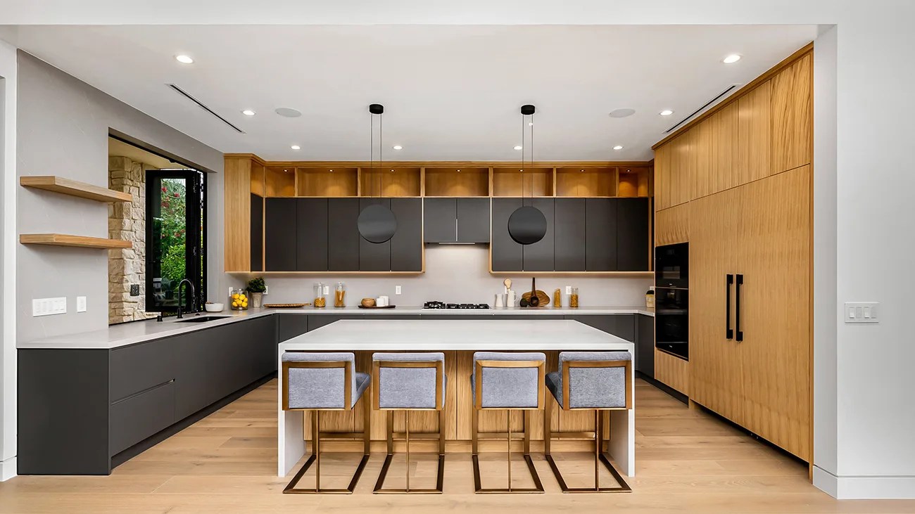 Modern kitchen with black and wood cabinets, white island countertop, four gray cushioned bar stools, and pendant lights.