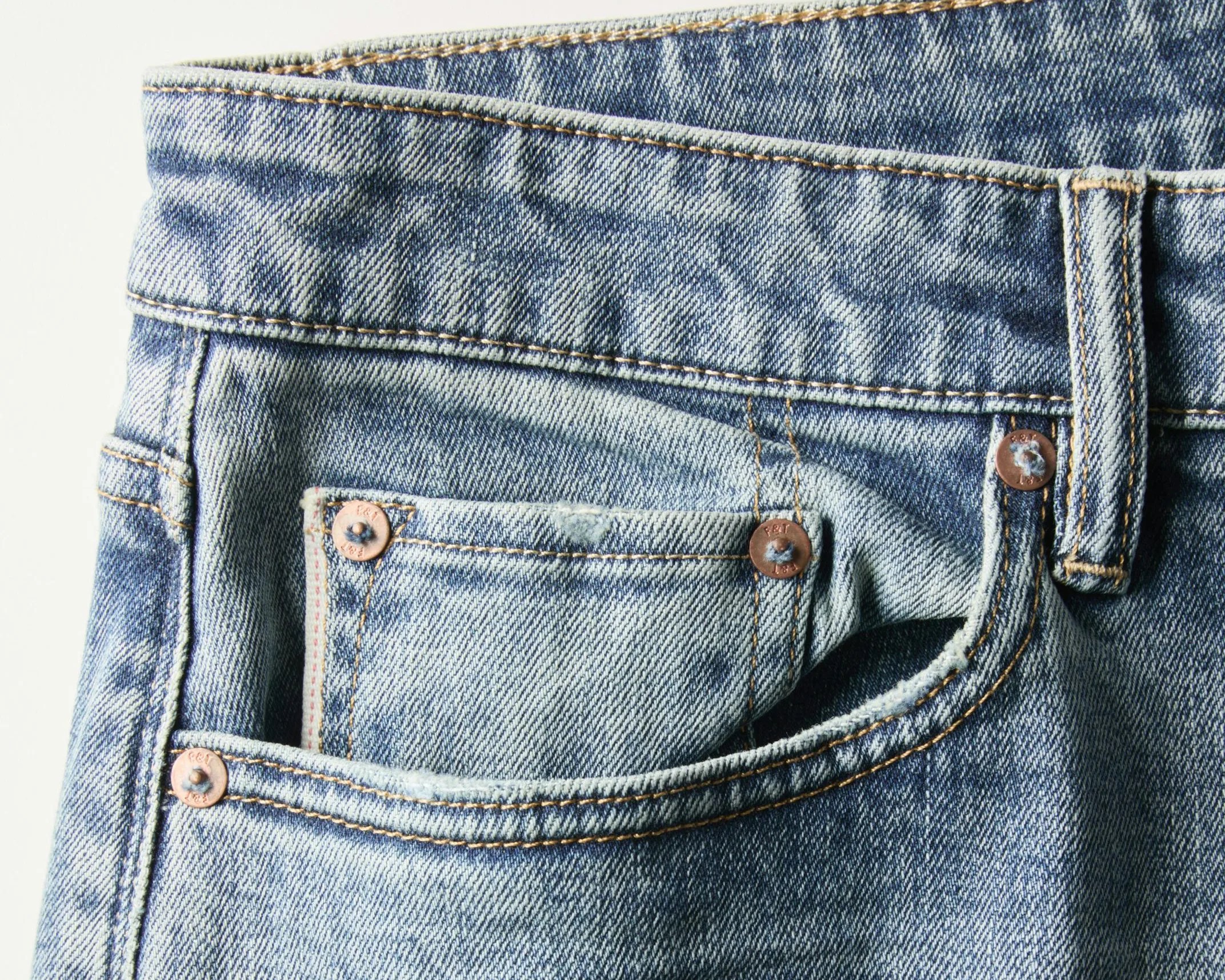 Close-up of light blue denim jeans waistband and front pocket with copper rivets and orange stitching.