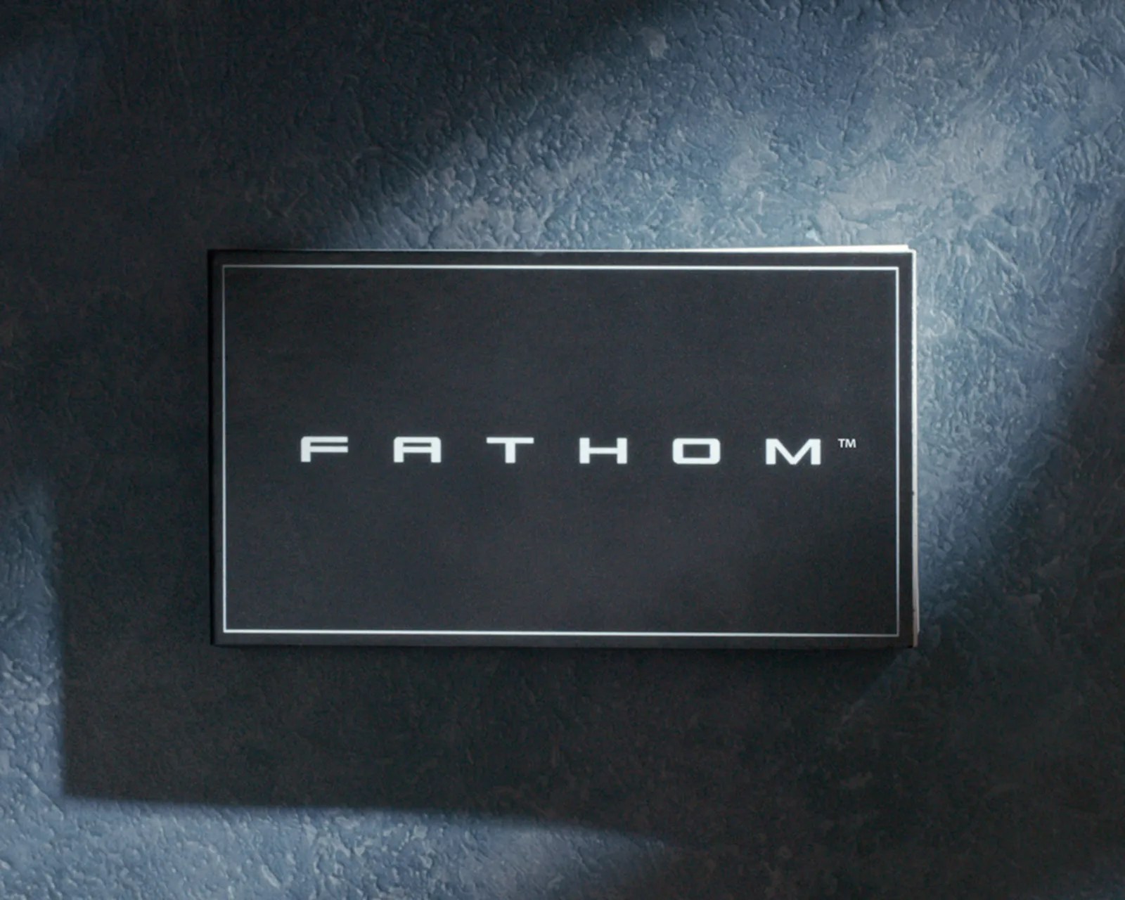Black rectangular box with the word "FATHOM" in white capital letters centered on a textured blue surface.