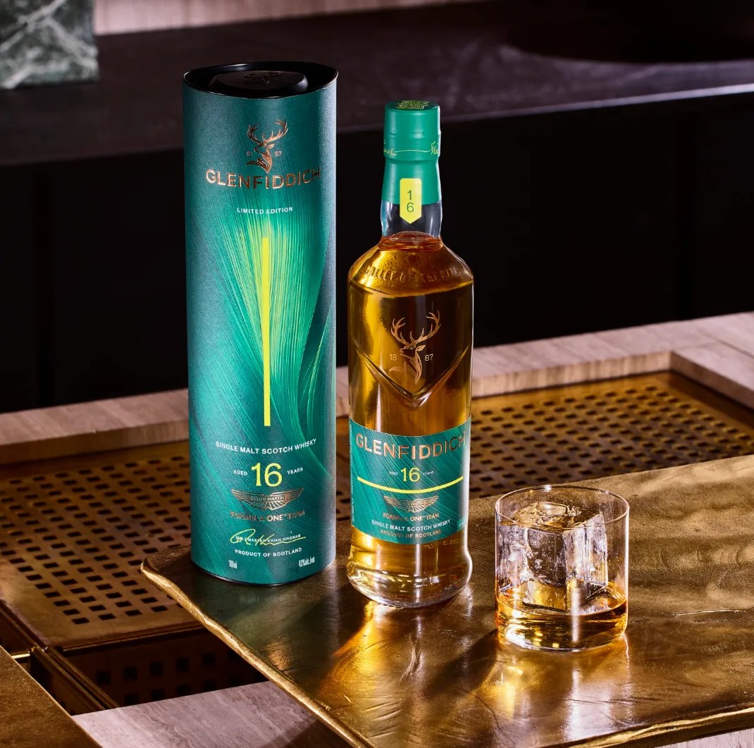 Bottle and cylindrical box of Glenfiddich 16-year-old single malt Scotch whisky next to a glass with whisky and ice on a brass tray.