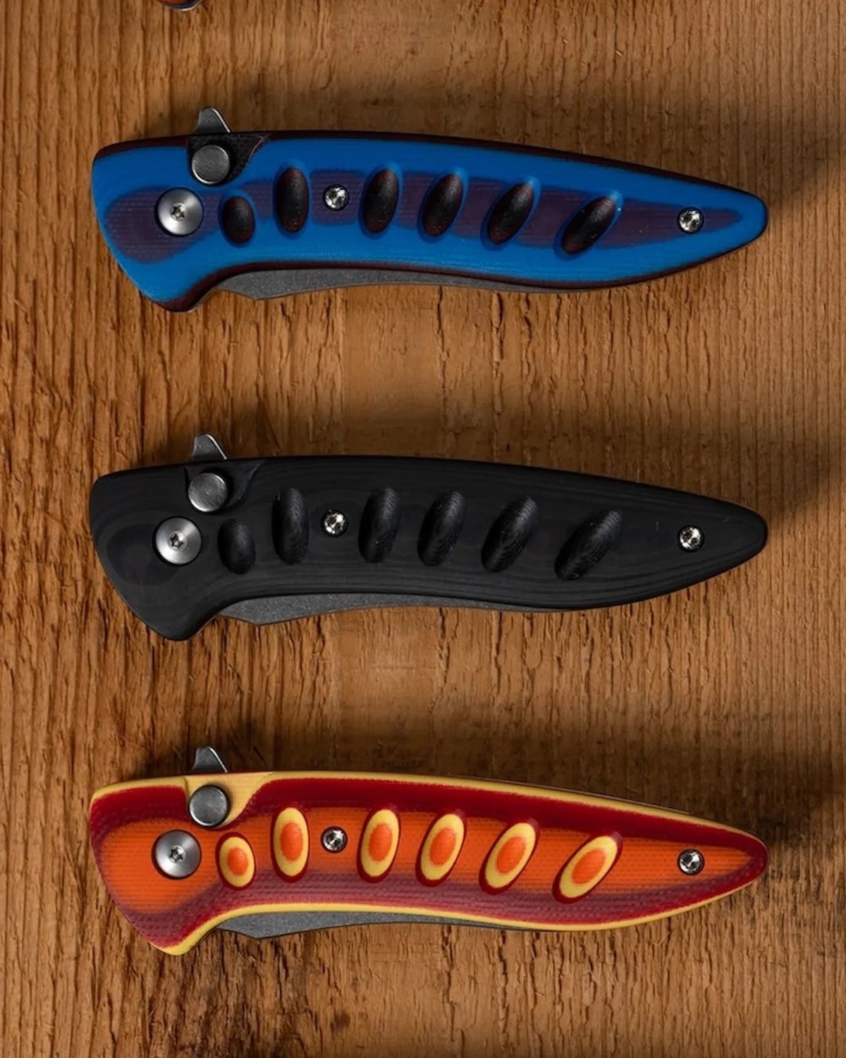Three closed folding knives with textured handles in blue, black, and orange-red colors on a wooden surface.