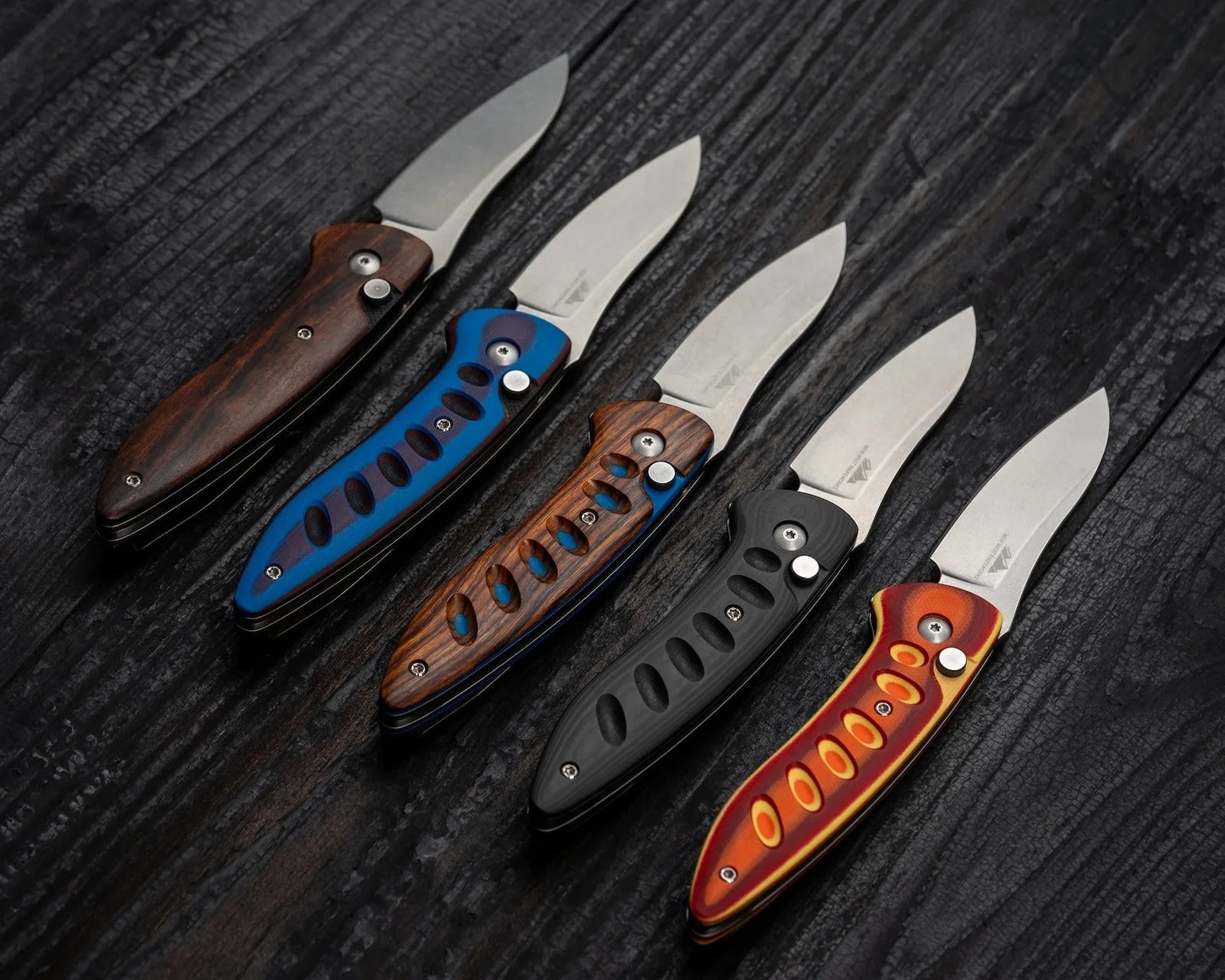 Five folding knives with partially serrated silver blades and colorful handles in wood, blue, black, and red-orange, arranged on a dark textured surface.