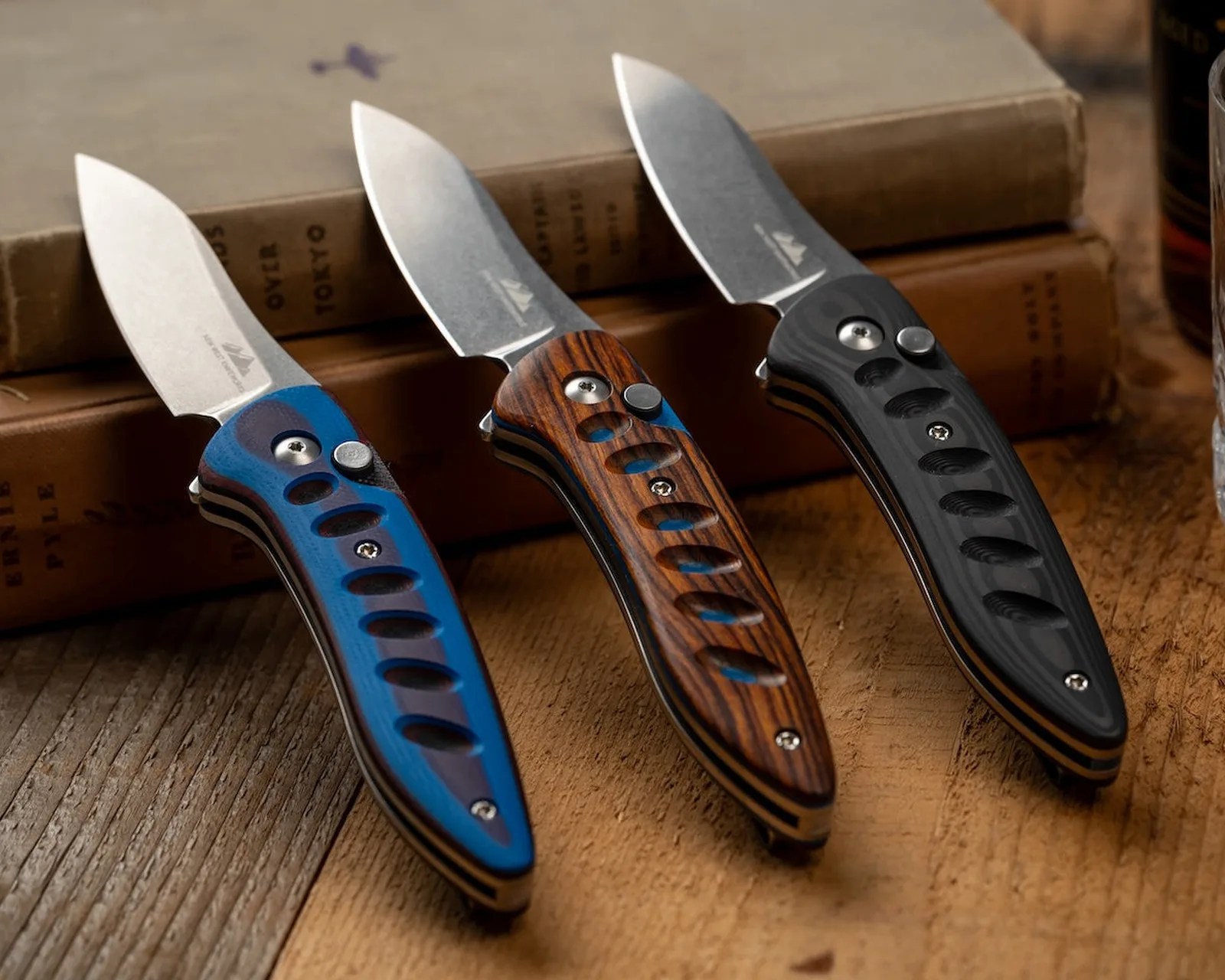 Three folding knives with stonewashed blades and textured handles in blue, wood grain, and black, resting on a wooden surface.