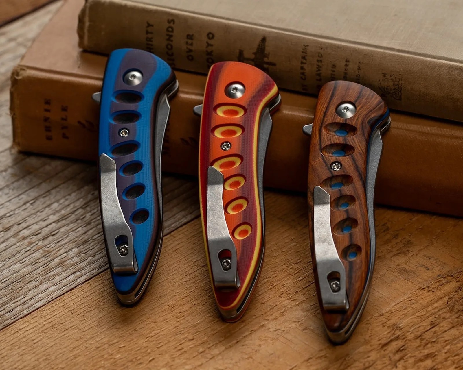 Three closed folding knives with blue, orange, and wood-grain handles resting on a wooden surface near two stacked books.