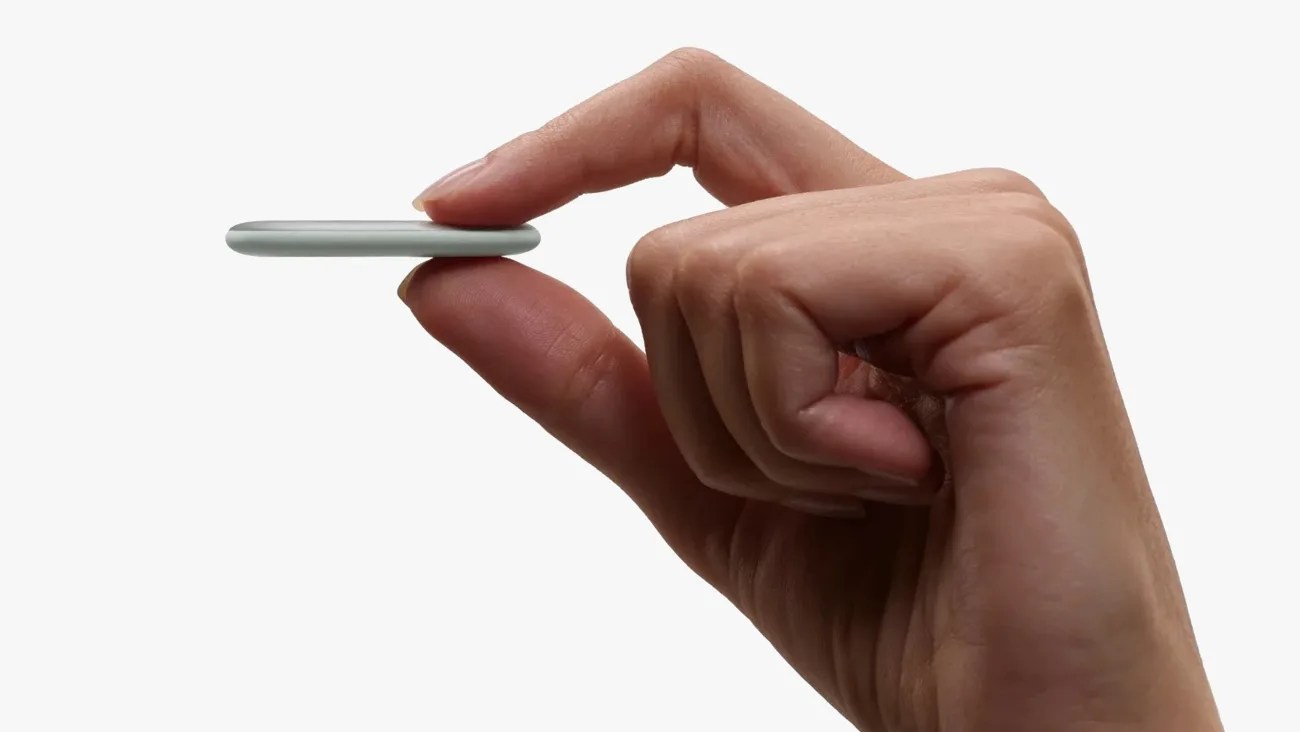 Hand holding a thin, round, silver-gray device between thumb and index finger against a white background.