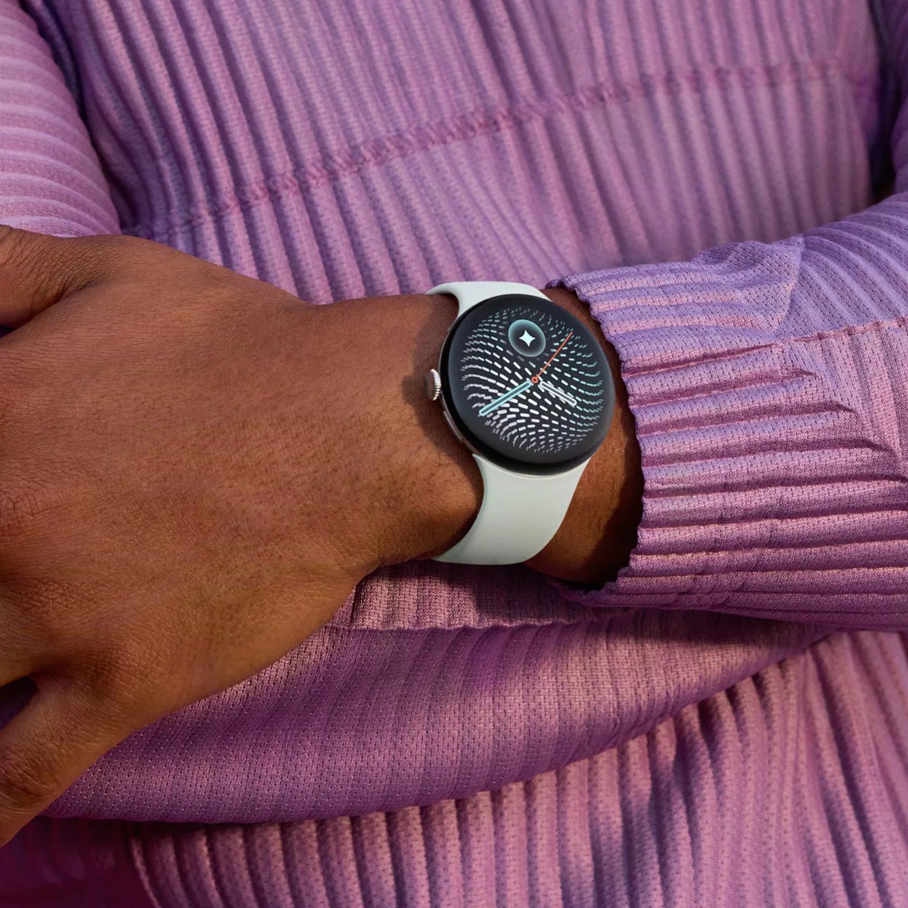 Smartwatch with a white band and black round face displaying a patterned watch face, worn on a wrist over a purple ribbed long sleeve.