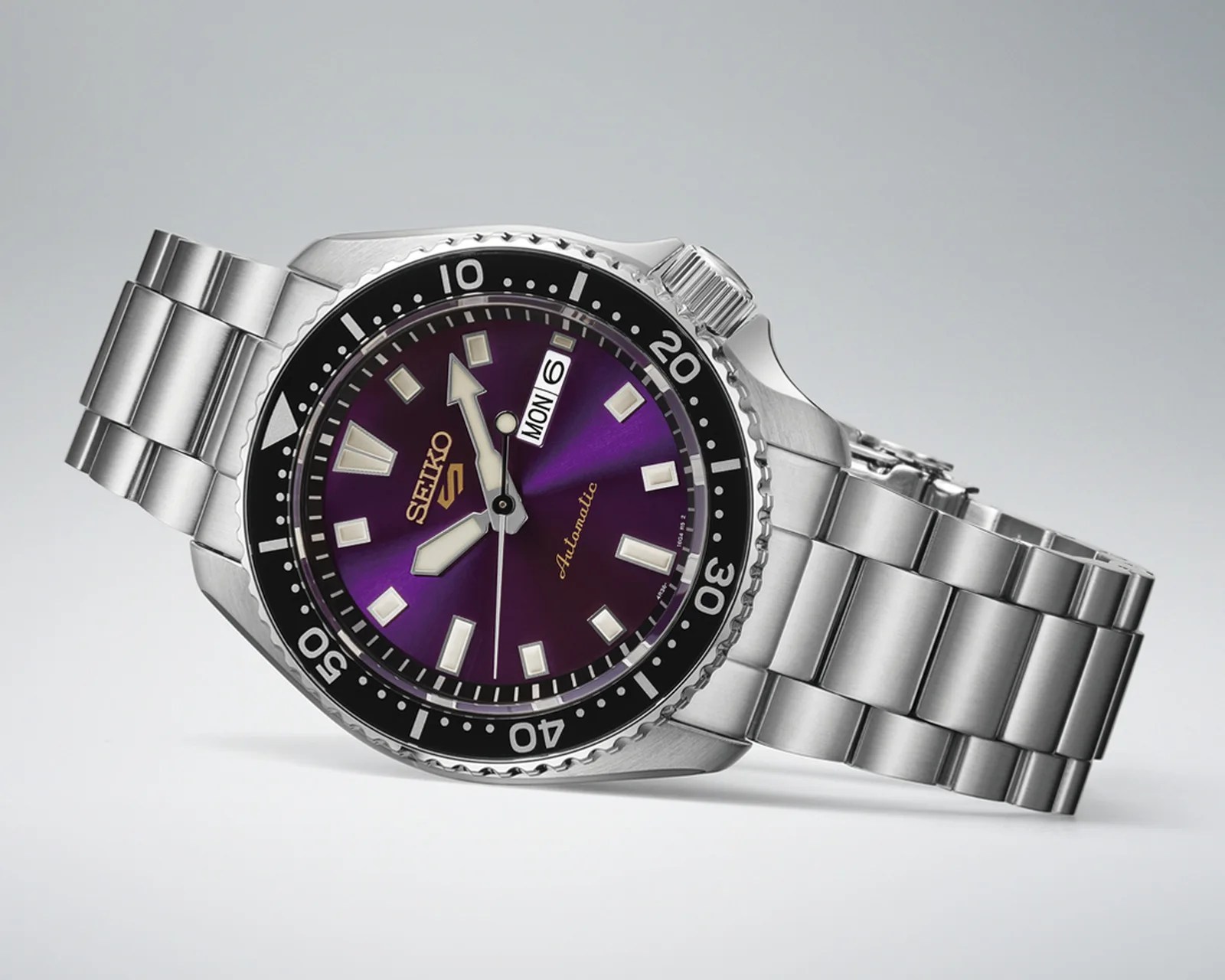 Seiko 5 automatic wristwatch with a purple dial, black bezel, and stainless steel bracelet.