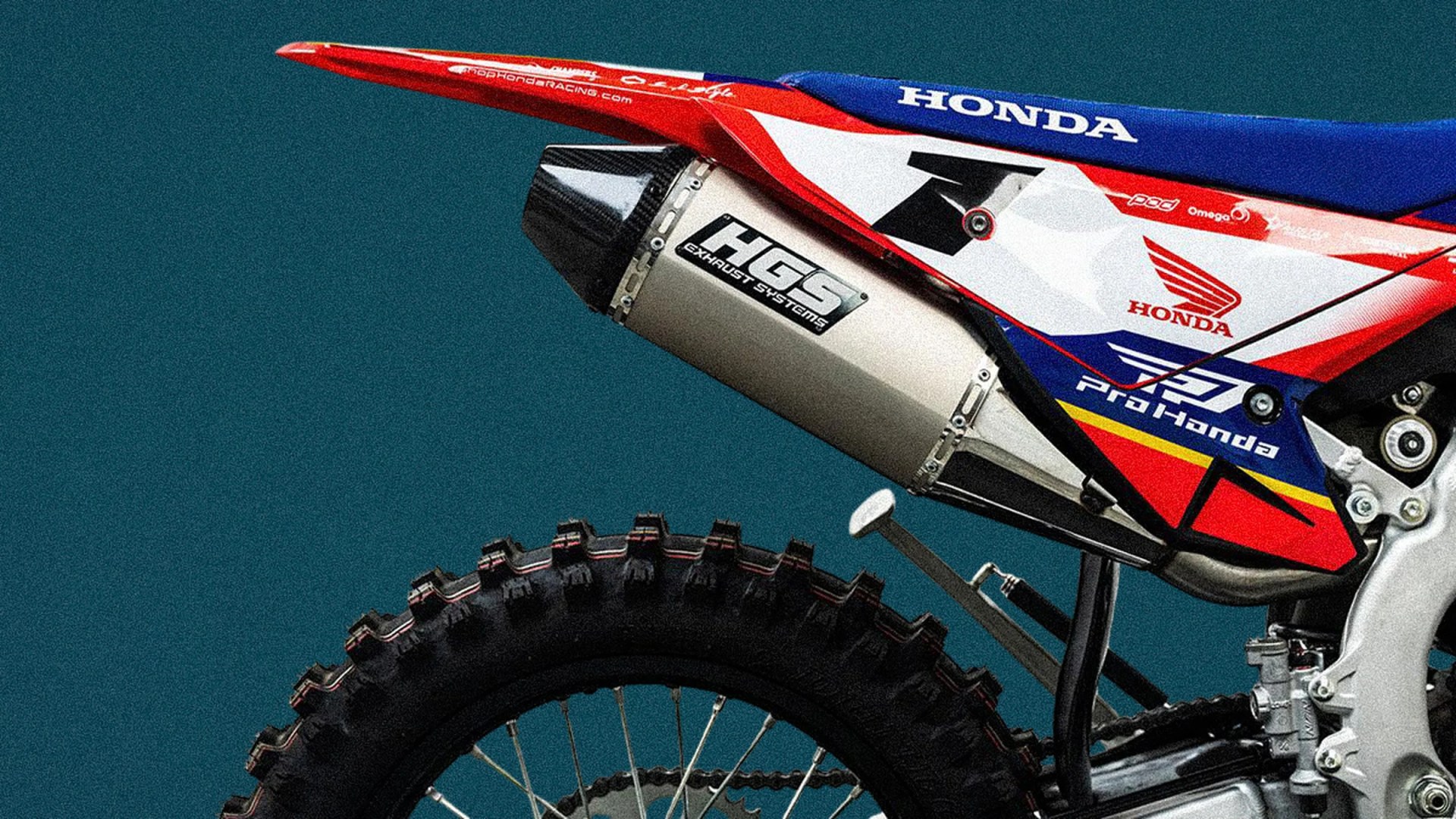 Rear section of a red, white, and blue Honda dirt bike with HGS exhaust system and knobby rear tire.