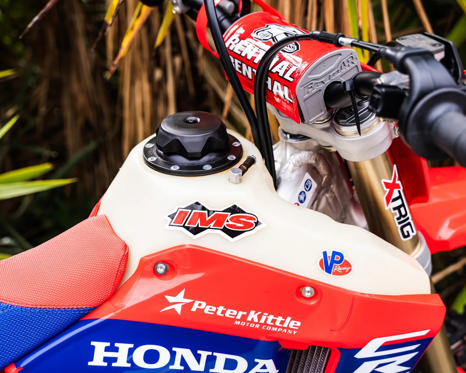 Close-up of a red, white, and blue Honda dirt bike fuel tank with IMS and VP Racing stickers and Renthal handlebar pad.