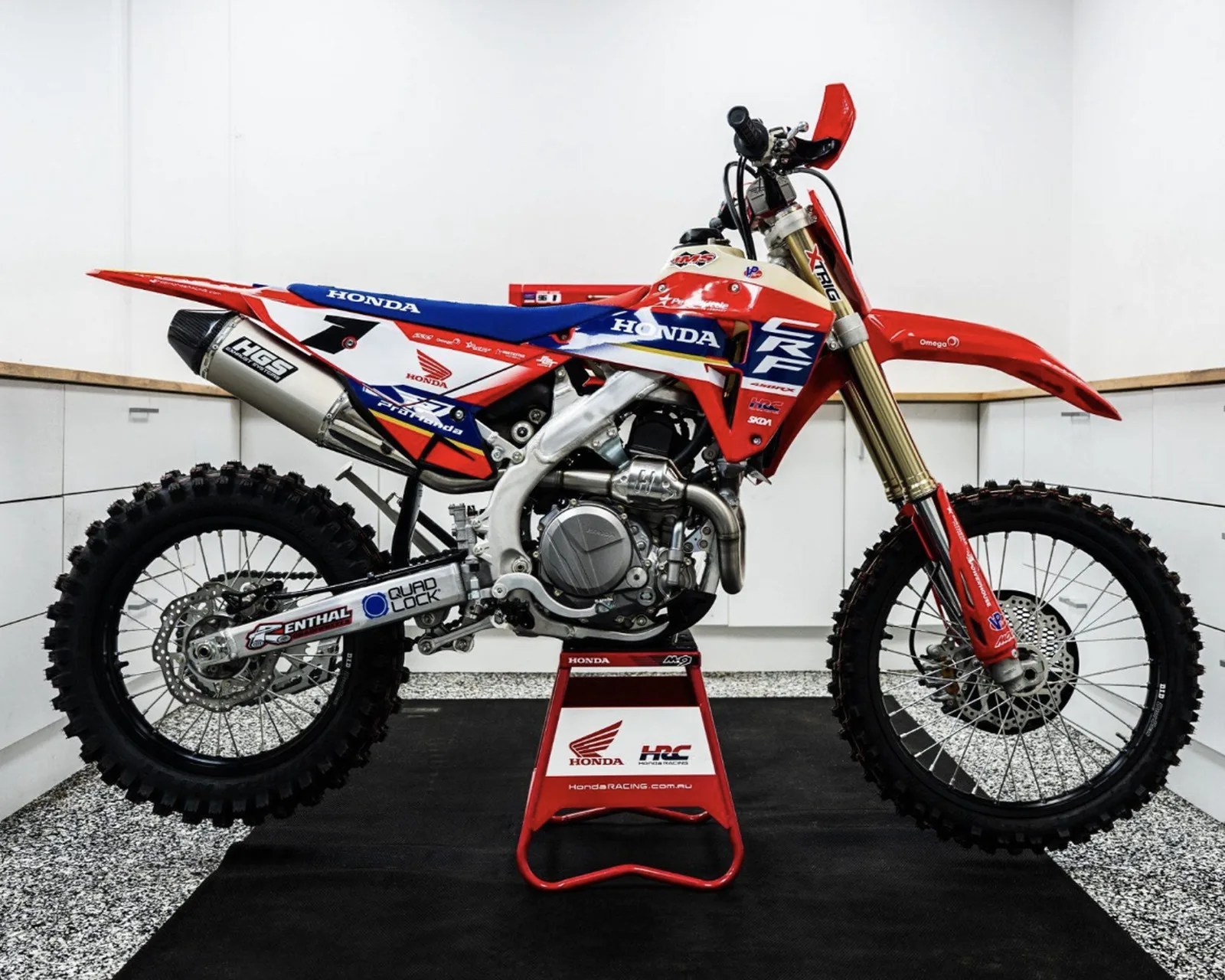 Red, white, and blue Honda CRF dirt bike with knobby tires on a red stand in a white garage.