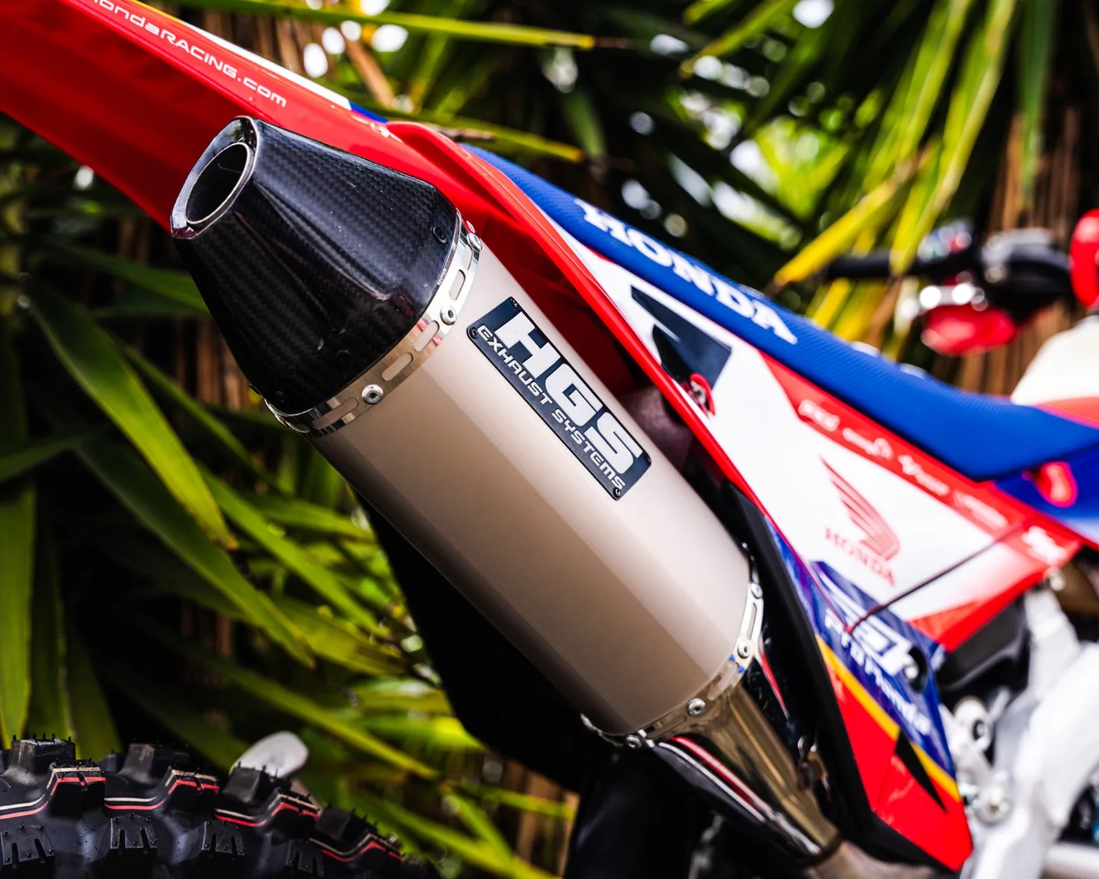 Close-up of a silver HGS exhaust system with a carbon fiber tip on a red, white, and blue Honda dirt bike.